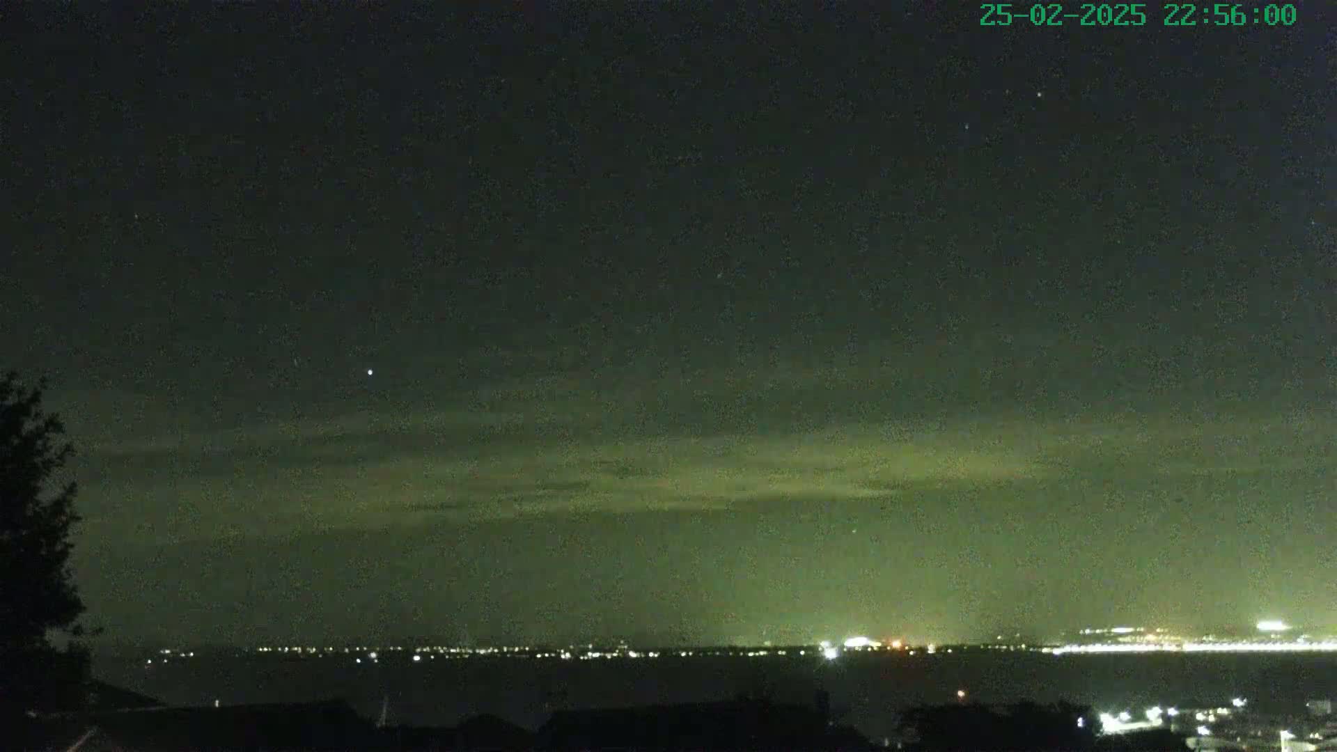 A nighttime view shows a body of water with city lights on the horizon under a dark sky with a few clouds and stars.
