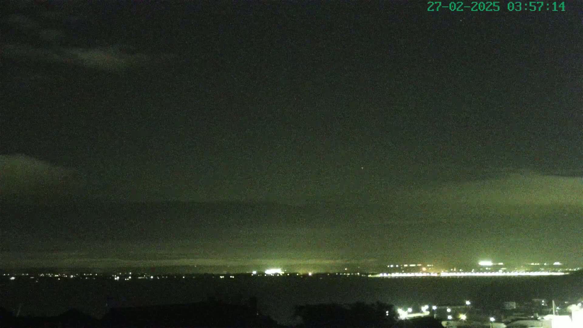 A nighttime, cloudy view of a distant city skyline illuminated by lights across a dark body of water.