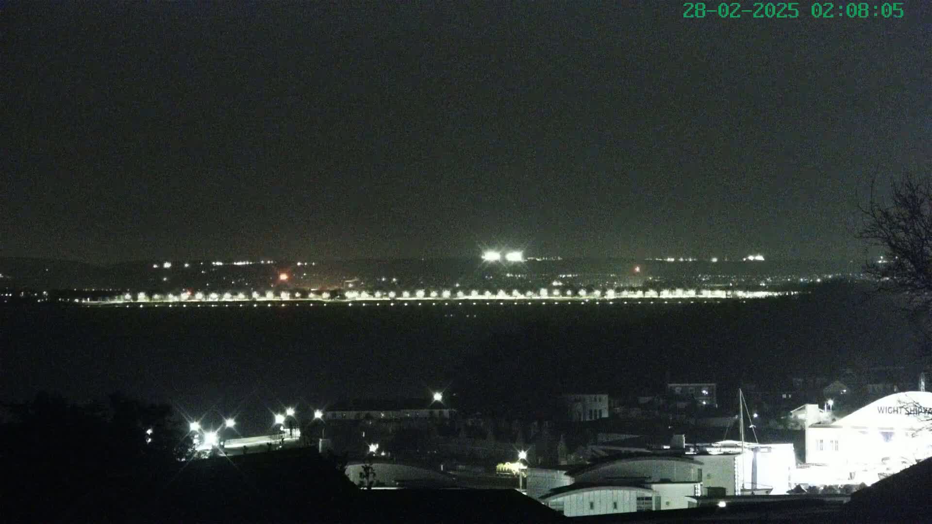 A nighttime view of a city skyline across a body of water, illuminated by streetlights and distant building lights under a dark sky.