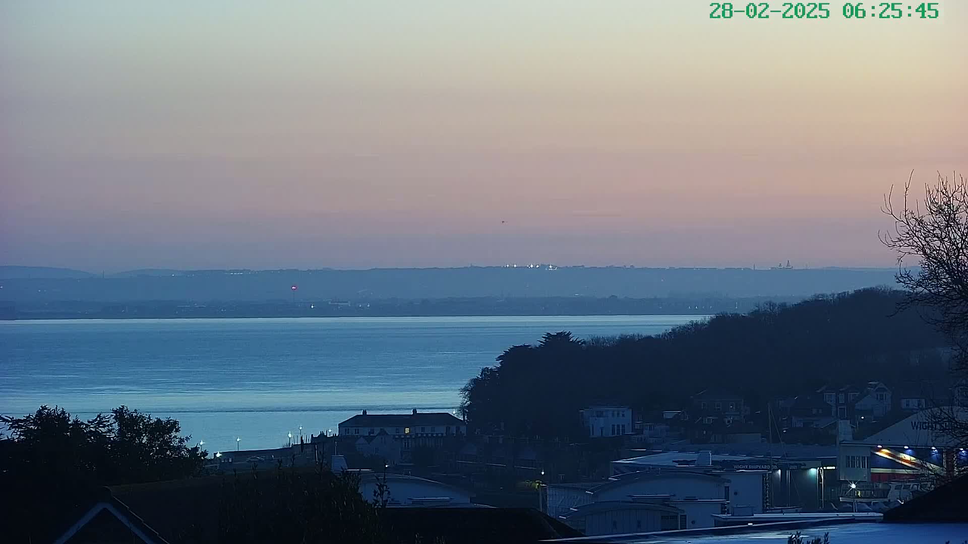 A twilight view shows a calm body of water, with a coastline of dark trees and buildings under a pale pink and purple sky.