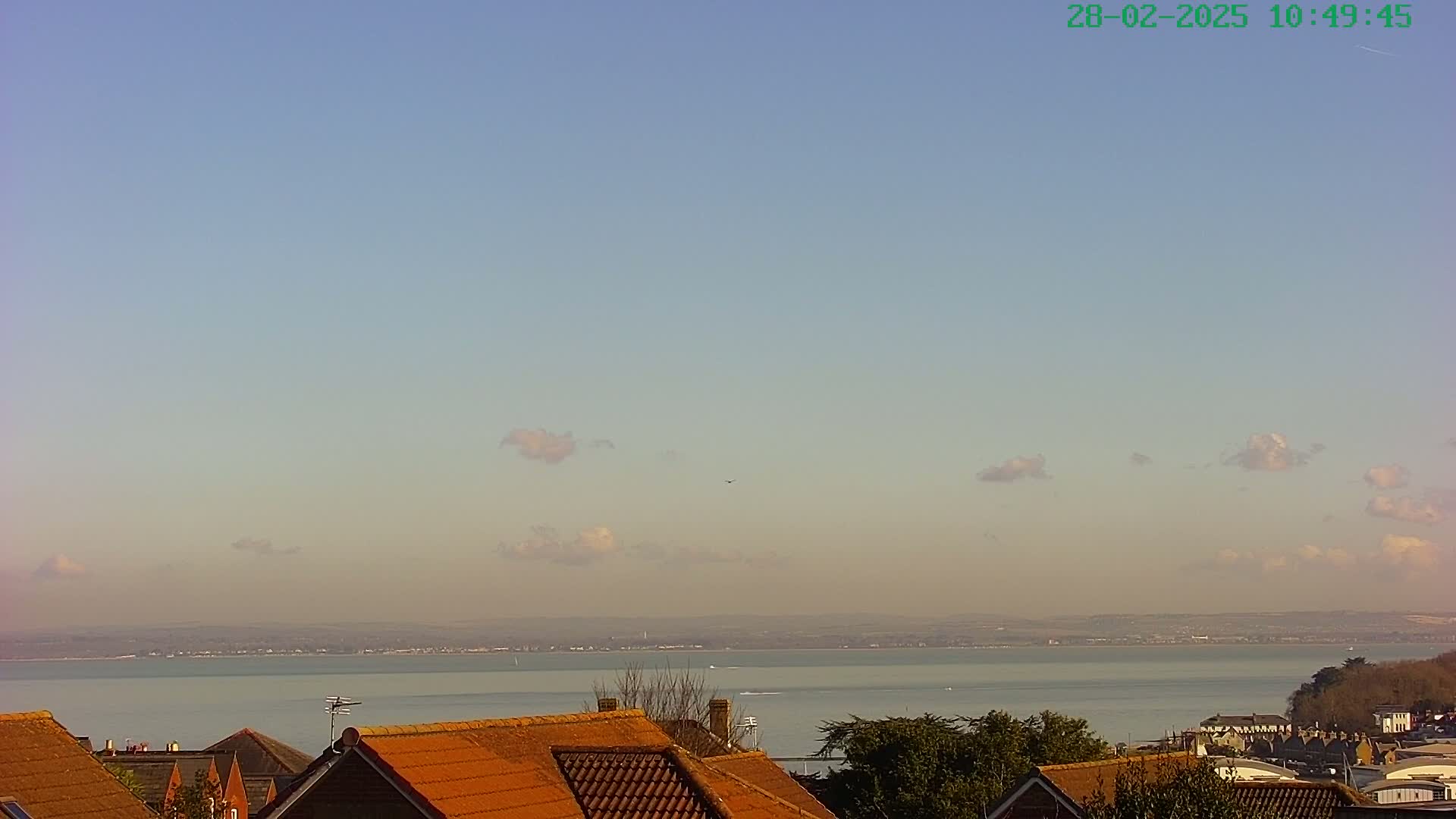 A mostly clear sky with a few scattered clouds overlooks a calm body of water and a distant shoreline, with orange-tiled rooftops in the foreground.