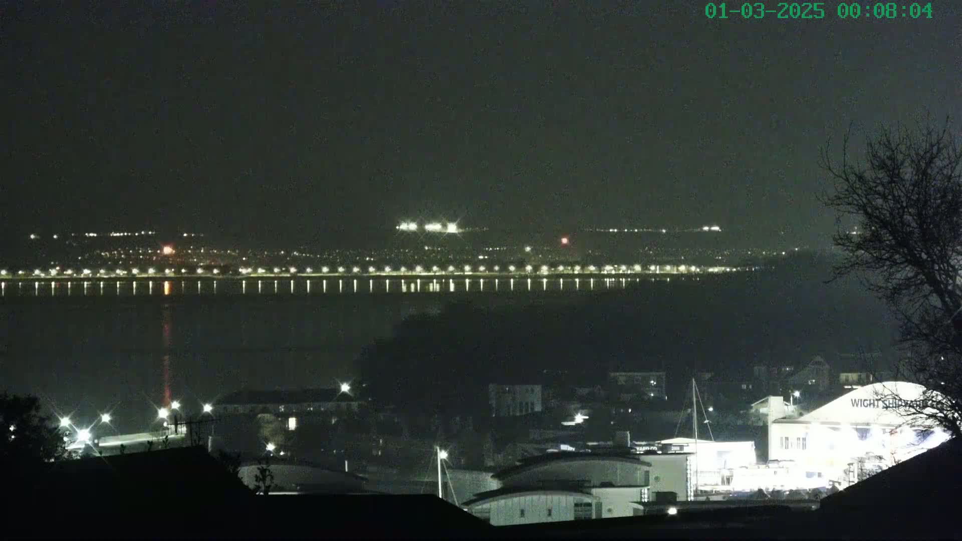 A nighttime view of a city across a body of water, with lights illuminating the waterfront and buildings, under a dark, clear sky.
