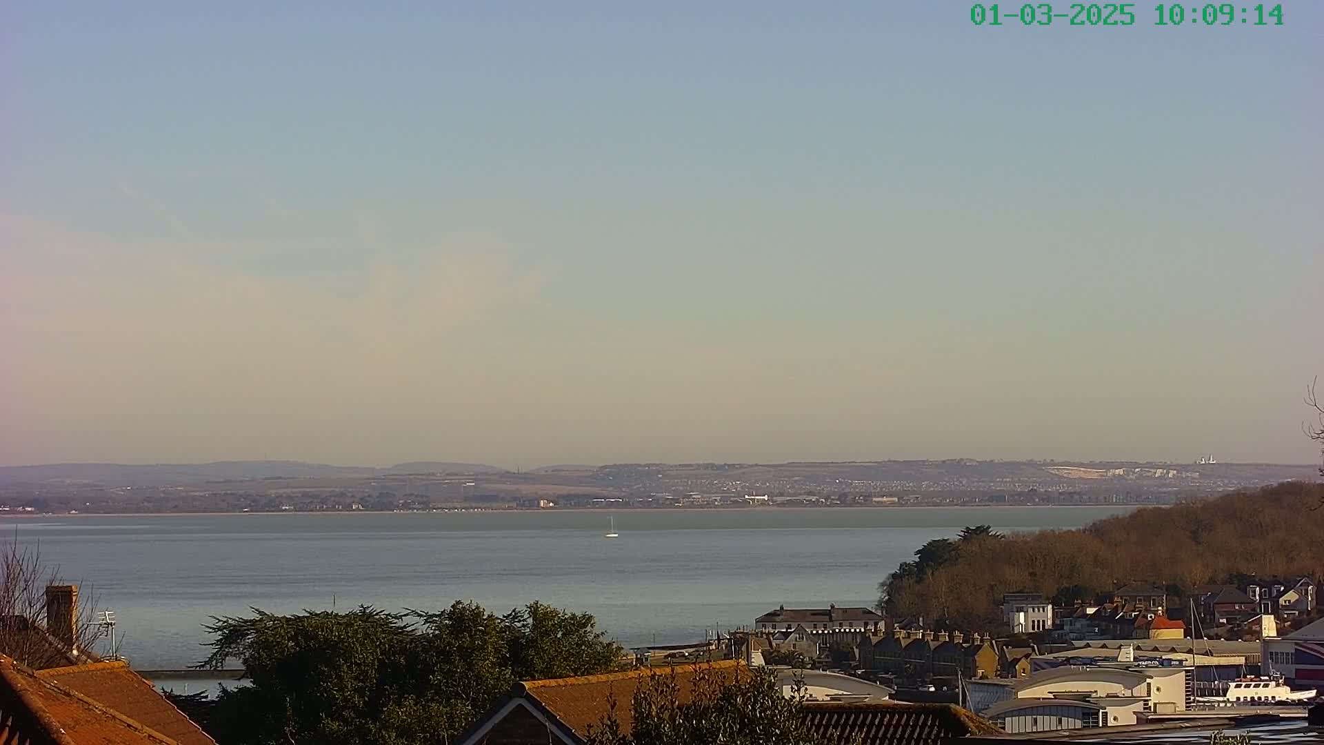 A mostly clear, sunny day overlooks a calm body of water with a distant town and hills visible in the background, partially obscured by haze, and rooftops and trees in the foreground.