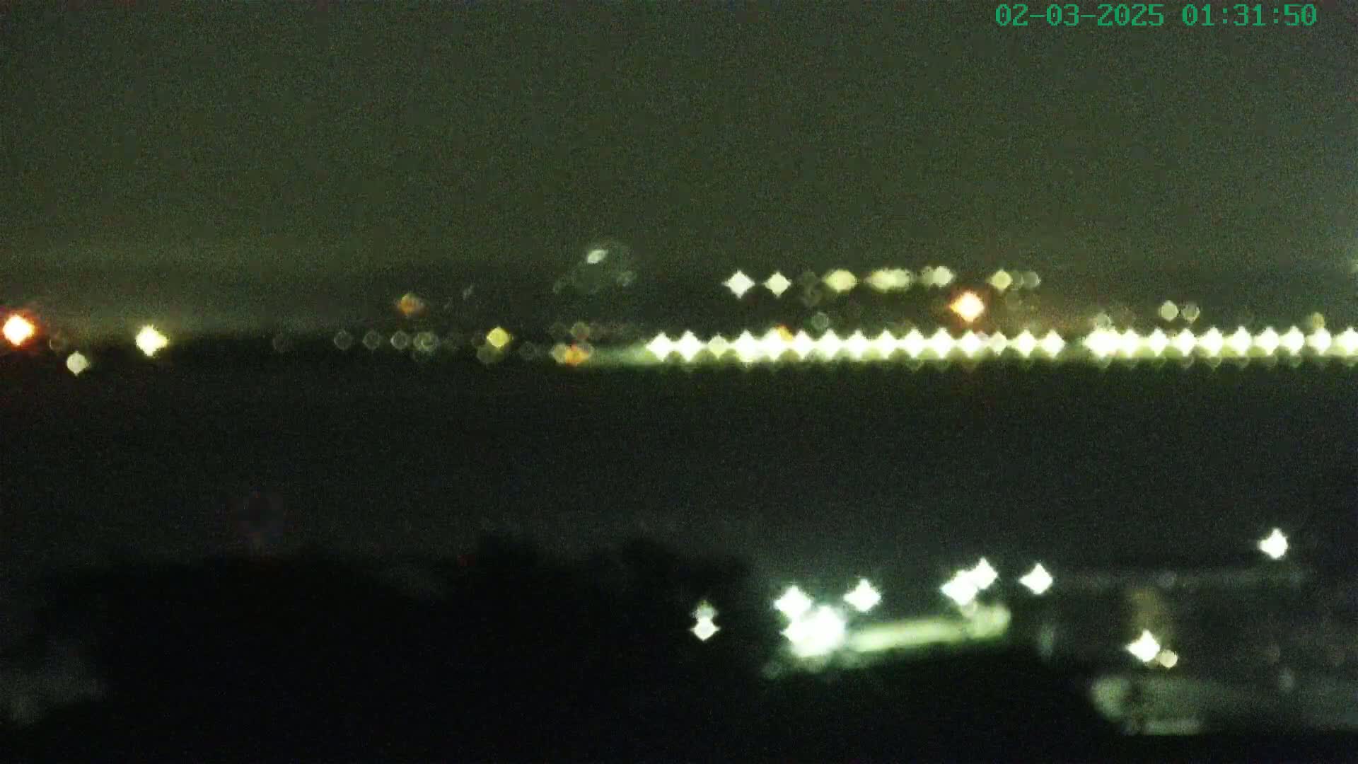 A blurry nighttime image shows a line of lights across a body of water, with more lights visible in the distance and closer to the bottom of the frame.