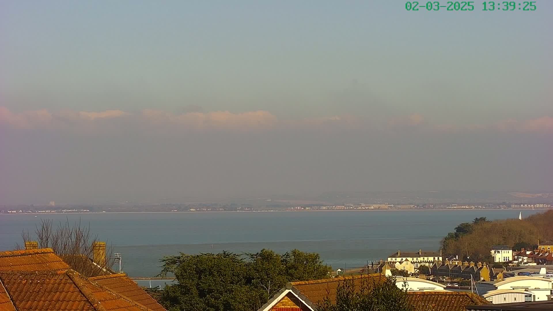 A hazy, sunny day reveals a calm body of water stretching to a distant coastline, viewed from behind a row of houses with reddish-brown roofs.