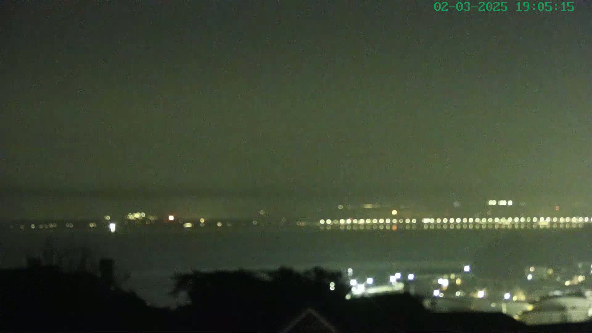 A nighttime long shot shows a hazy body of water with a line of lights on the far shore, and a closer collection of lights on the lower right.