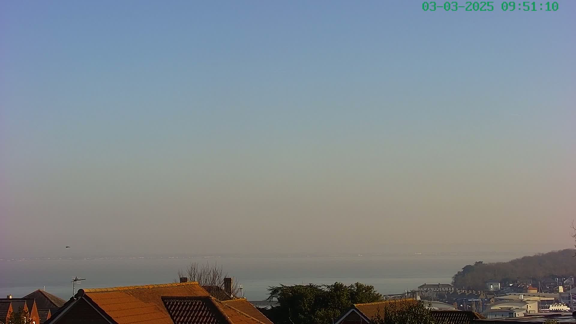 A hazy view of a calm body of water extends to the horizon behind the reddish-brown rooftops of houses and some trees.