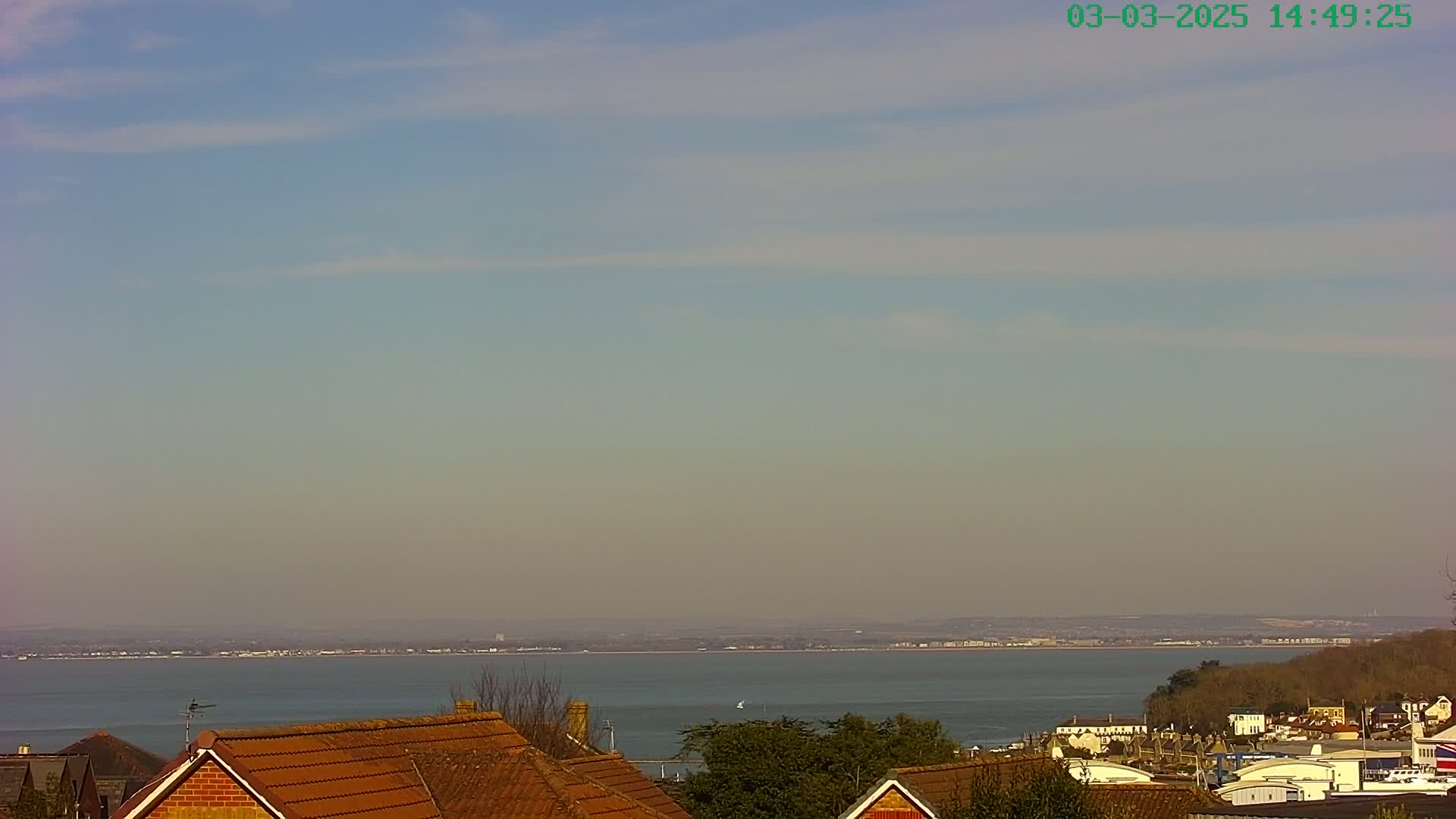 A hazy, sunny day overlooks a calm body of water with a distant shoreline, visible behind rows of residential rooftops.