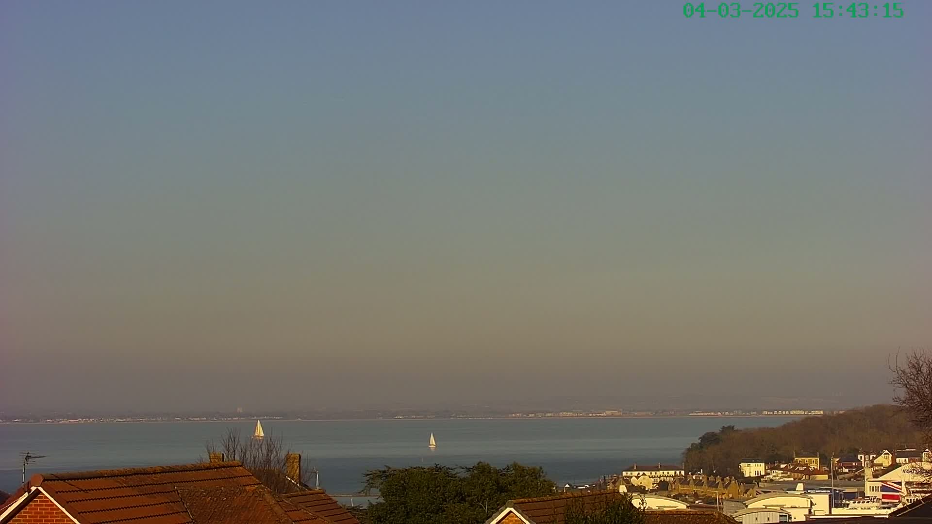 A hazy, calm day overlooks a body of water with two sailboats, a distant shoreline, and rooftops in the foreground.