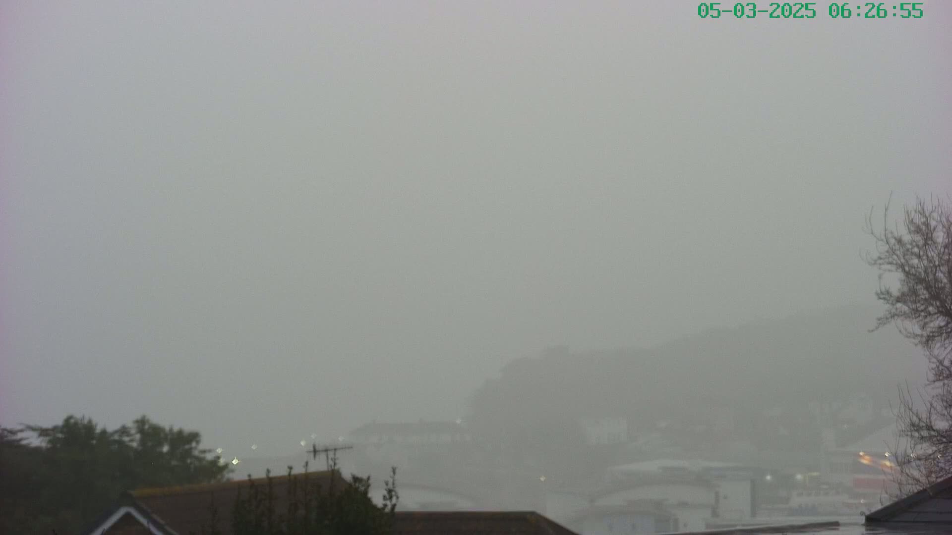 A foggy scene shows a distant town obscured by thick fog, with the tops of houses and trees visible in the foreground.