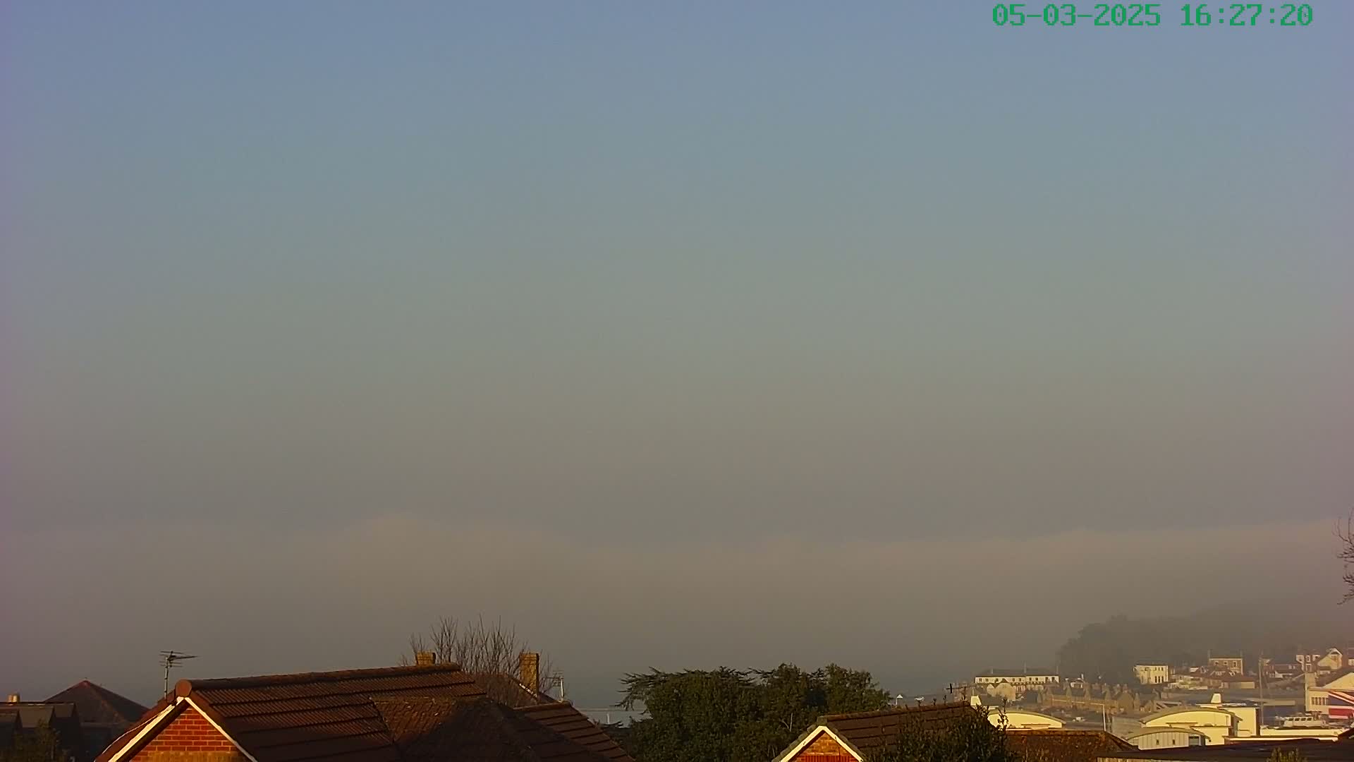 A hazy, low-lying fog obscures a distant town and coastline, partially visible behind the brown-tiled roofs of houses in the foreground.