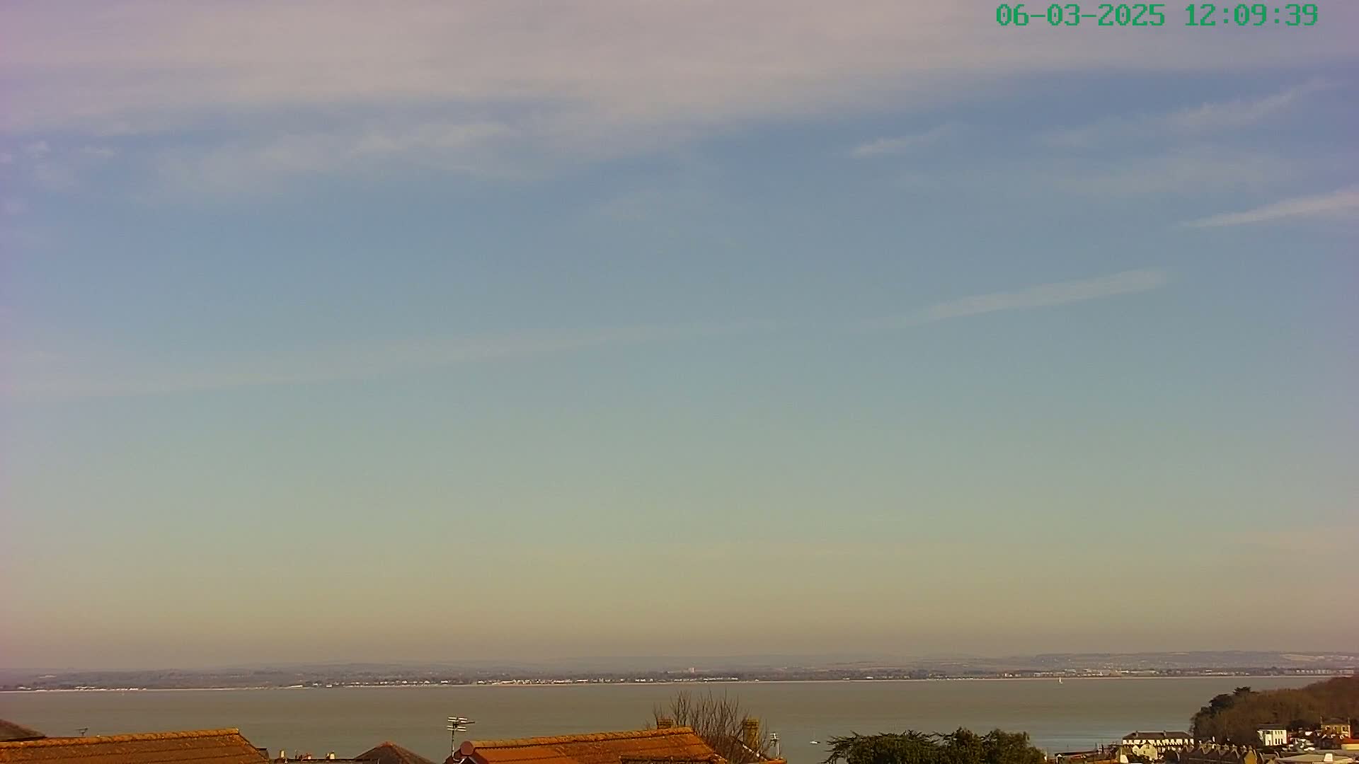 A calm, slightly hazy day reveals a wide expanse of water with a distant shoreline and a few houses with orange rooftops in the foreground.