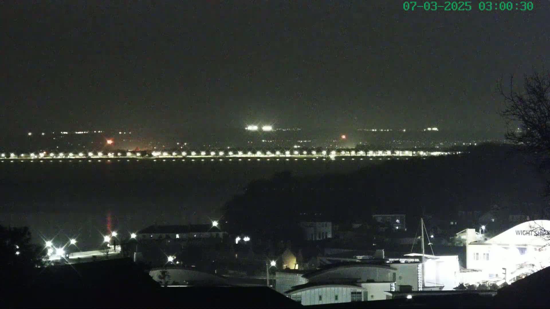 A nighttime view of a city skyline across a body of water, with scattered lights illuminating the foreground and background under a dark, clear sky.