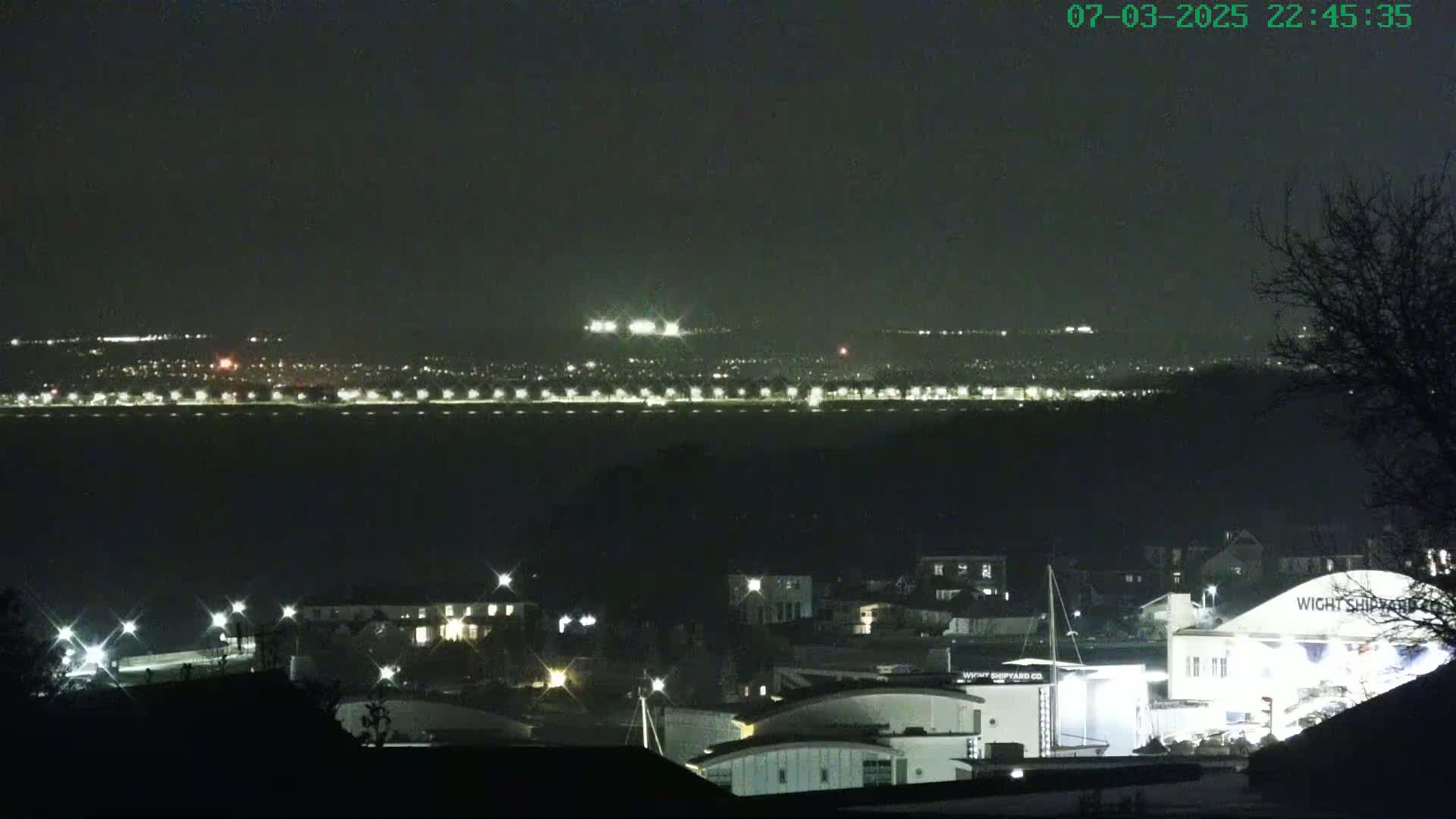 A nighttime long shot shows a city skyline across a dark body of water, with a closer view of buildings and lights along the waterfront.
