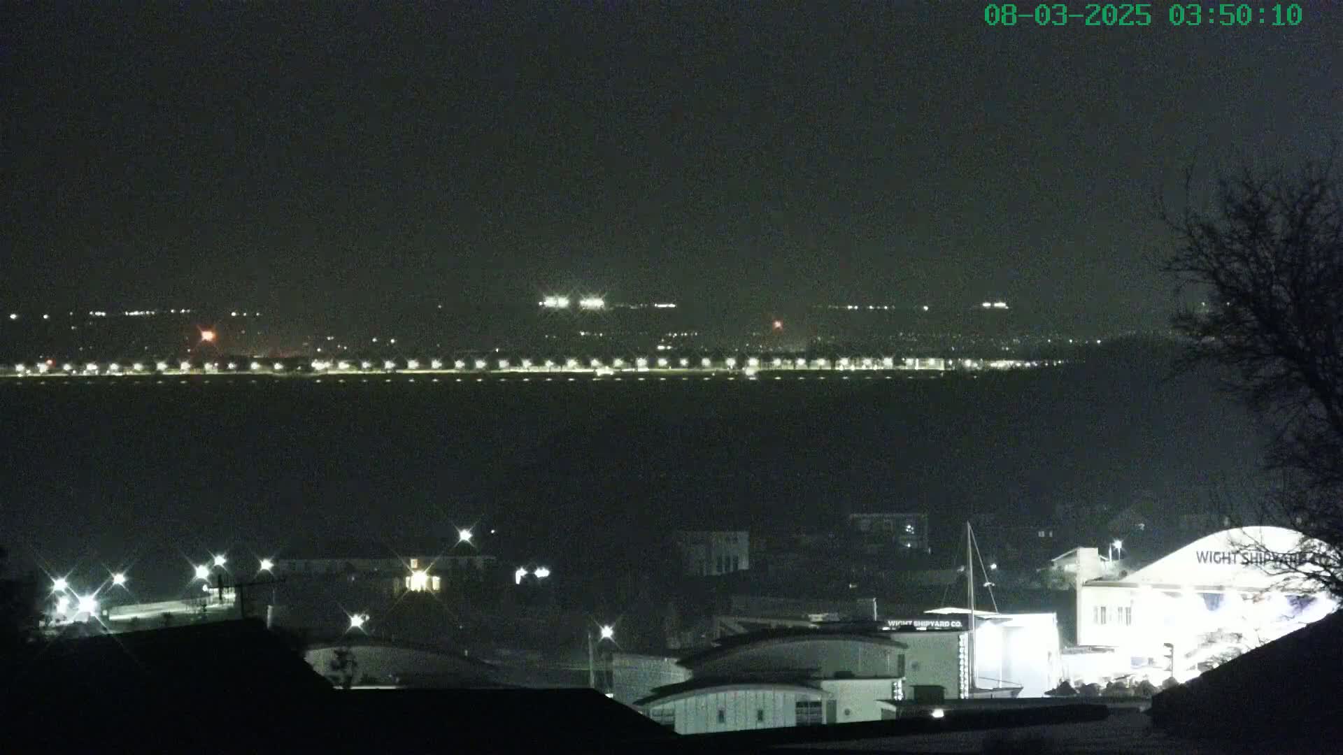 A nighttime, long shot of a waterfront area with a distant shoreline lit with lights across a dark body of water, and buildings and lights closer to the viewer.