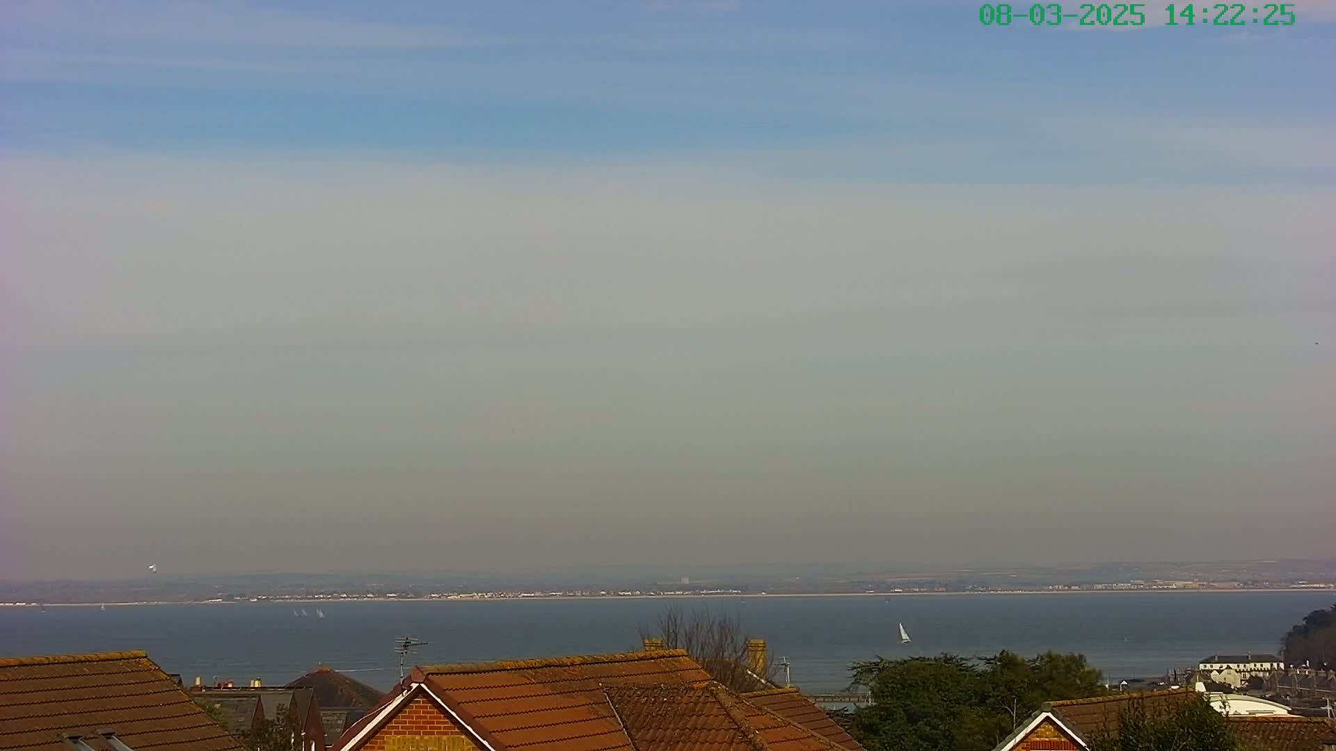 A hazy, sunny day overlooks a calm body of water with distant sailboats and rooftops in the foreground.