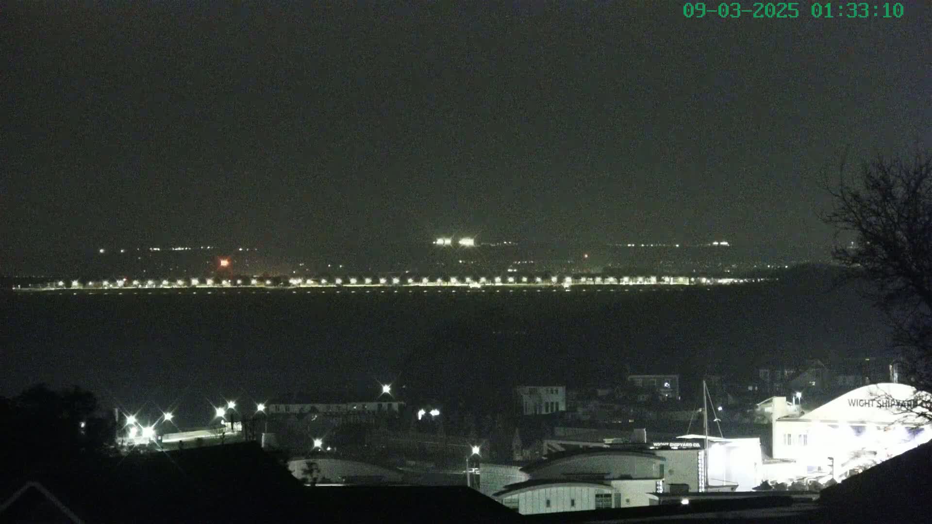 A nighttime view of a town lit by streetlights and buildings, across a body of water from a distant shoreline also showing lights.