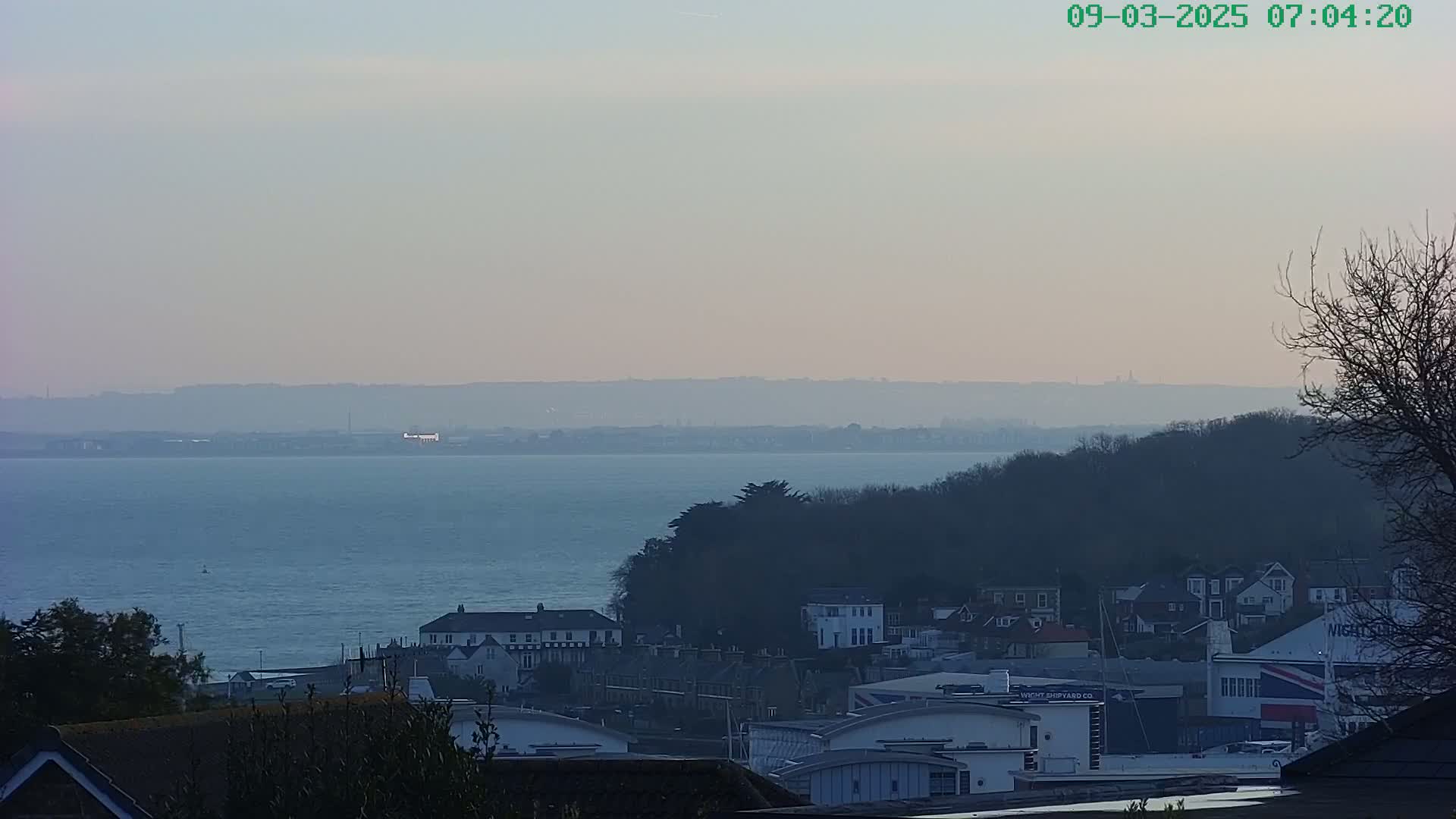 A hazy, pale-blue body of water stretches to a distant, indistinct shoreline, overlooking a collection of buildings nestled amongst trees on a hillside.