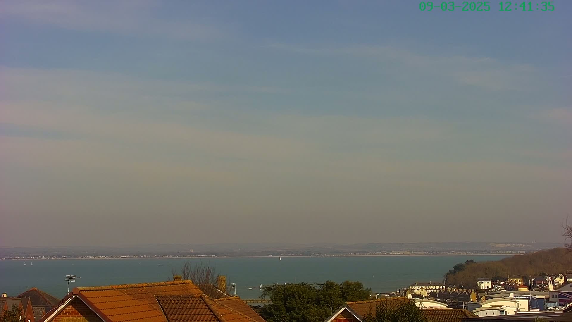 A hazy, sunny day overlooking a calm body of water with a distant shoreline, featuring rooftops of houses in the foreground.