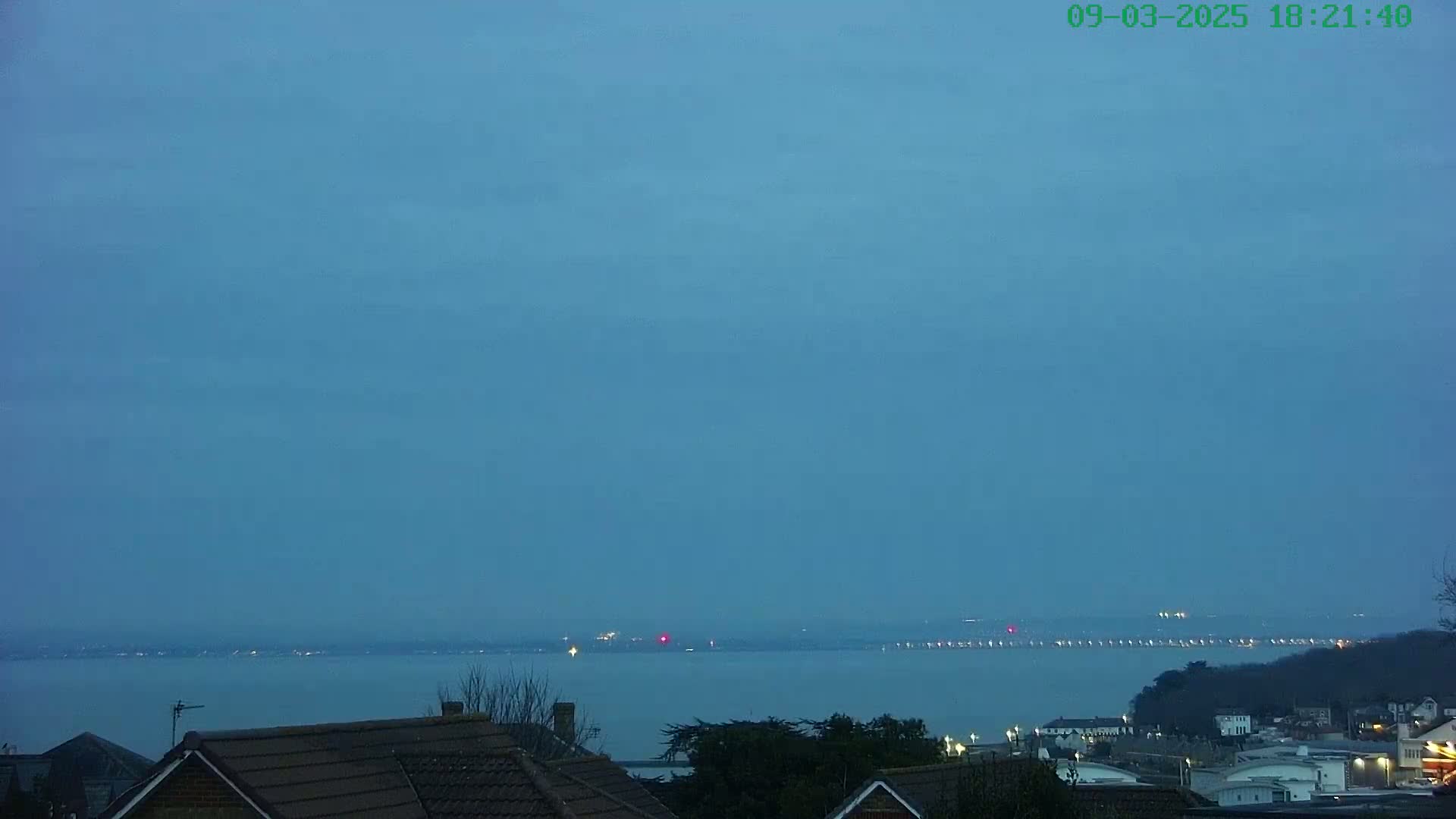 A twilight view shows a calm body of water extending to a distant shoreline with faint lights, viewed from behind houses with tiled roofs.