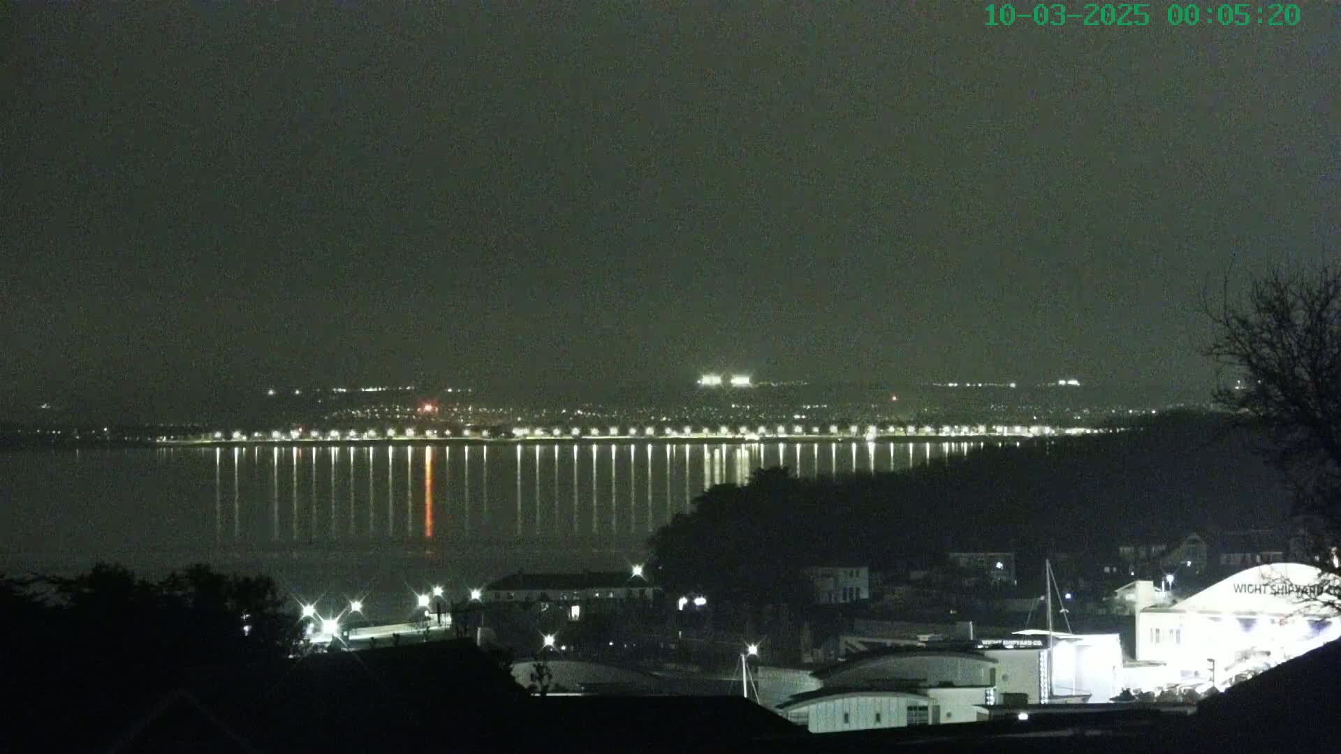 A nighttime view of a city across a calm body of water, illuminated by lights reflecting on the water's surface under a dark sky.