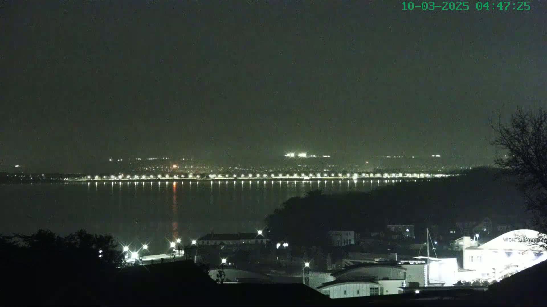 A nighttime view across a calm body of water shows a distant city skyline illuminated by lights, with a closer foreground of buildings and trees under a dark, hazy sky.