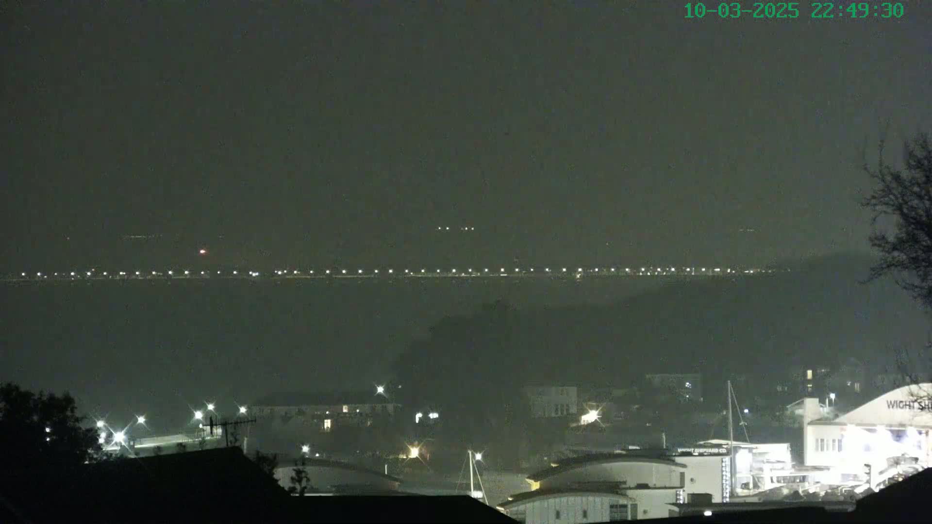 A nighttime view of a hazy waterfront, showing a distant line of lights across the water and a closer collection of buildings and boats under a dark sky.