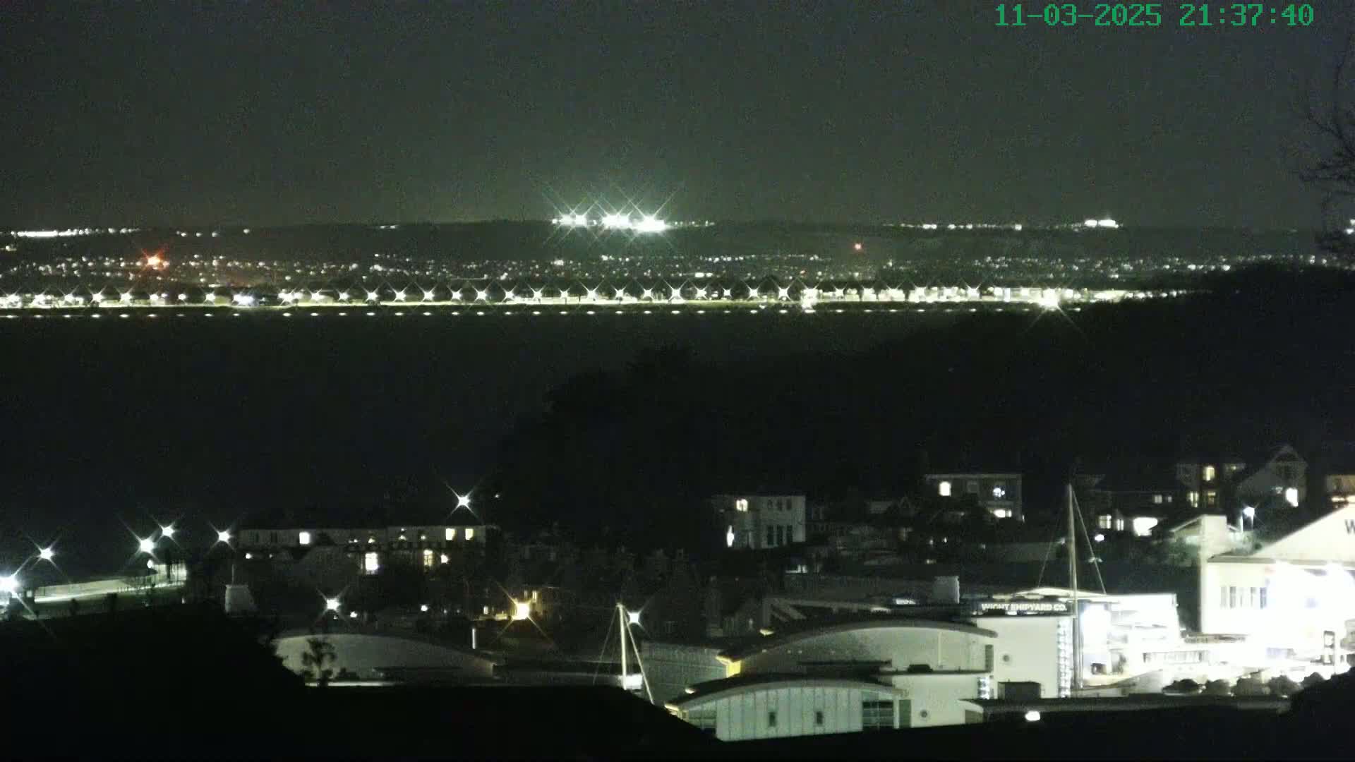 A nighttime view of a town situated near a body of water, with a distant city skyline illuminated under a clear, dark sky.