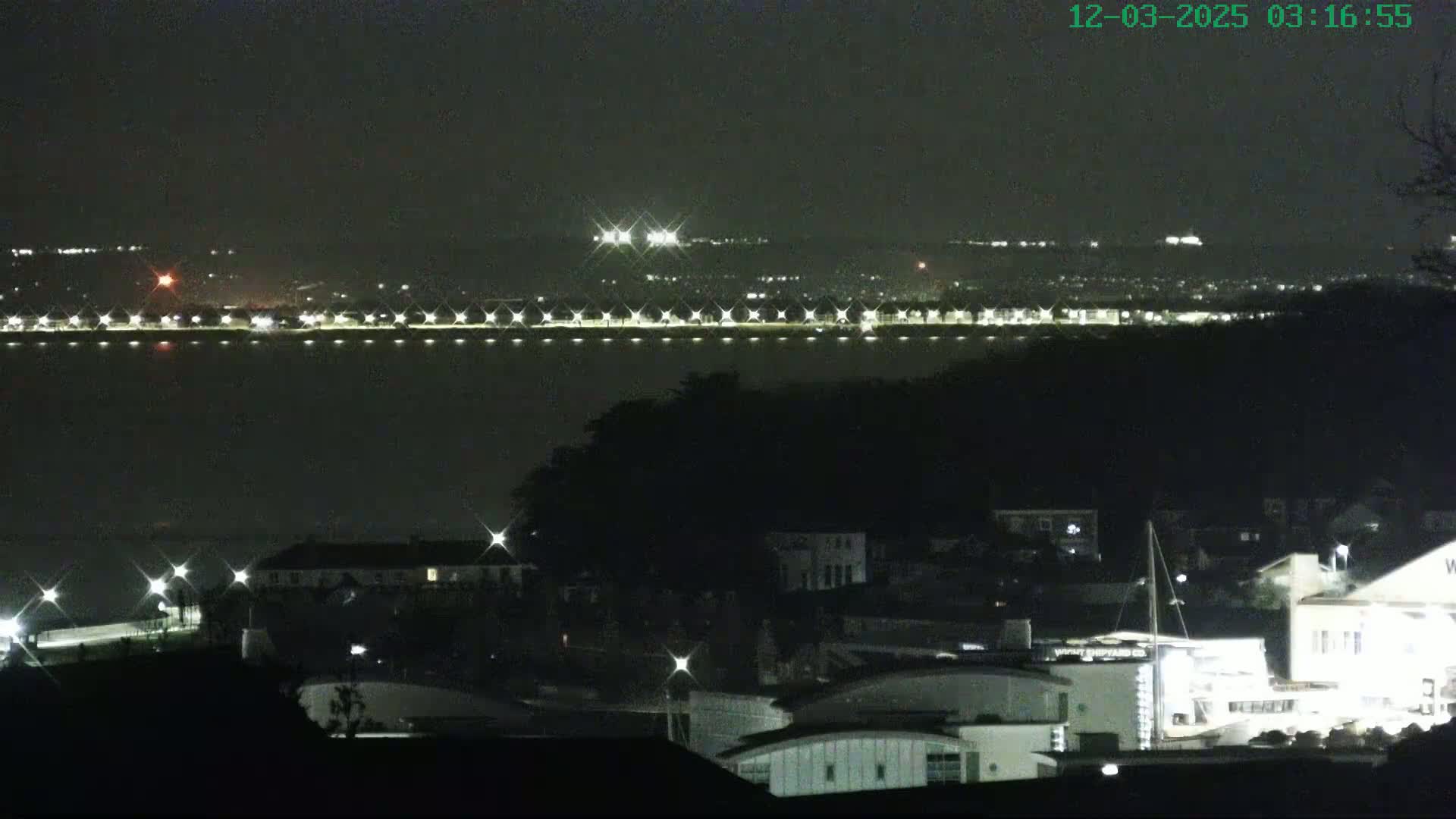 A nighttime view of a city across a body of water, with a line of lights along the waterfront and buildings silhouetted against the dark sky.