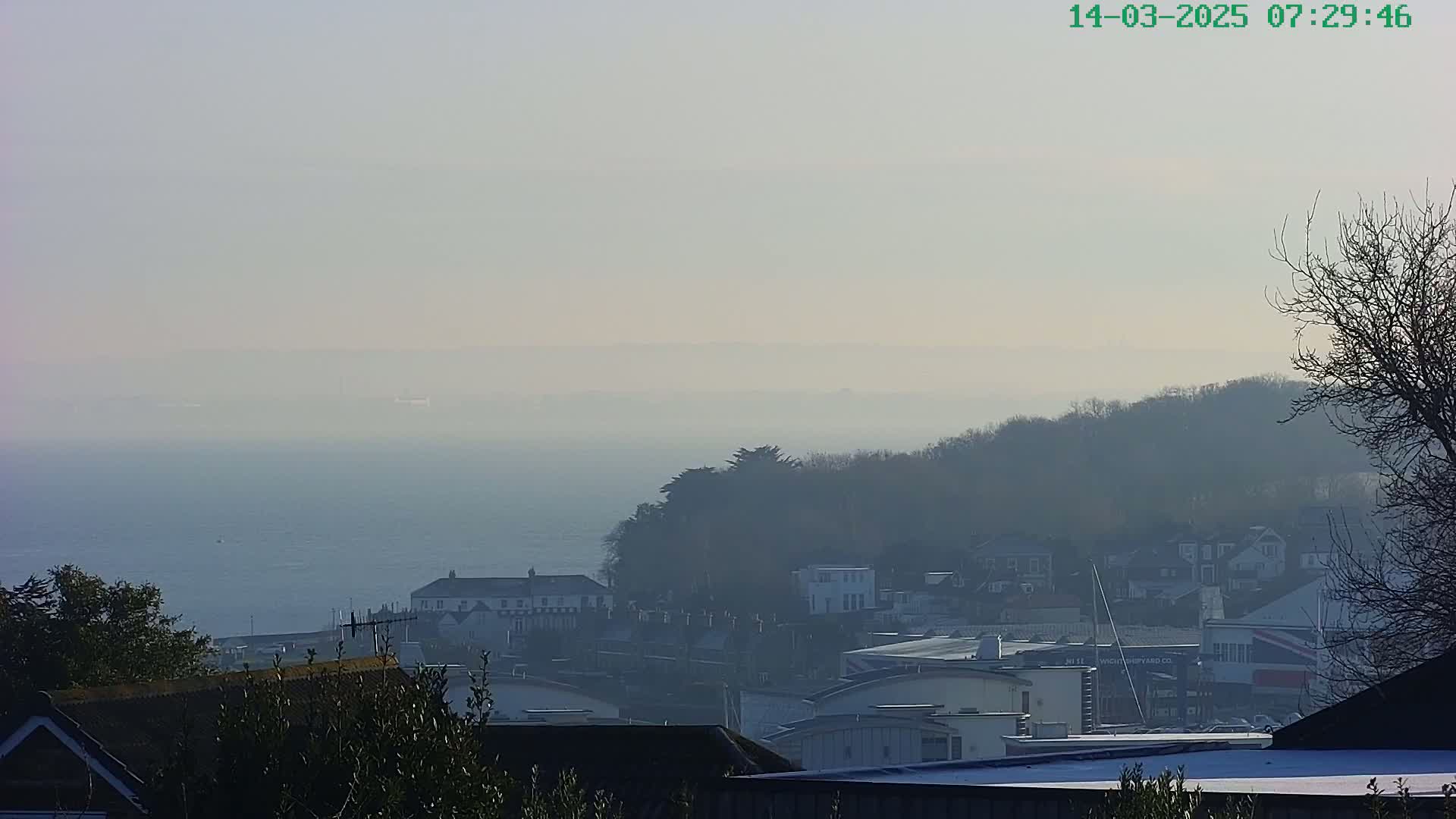 A hazy, distant view of a coastal town nestled amongst hills overlooking a calm body of water.