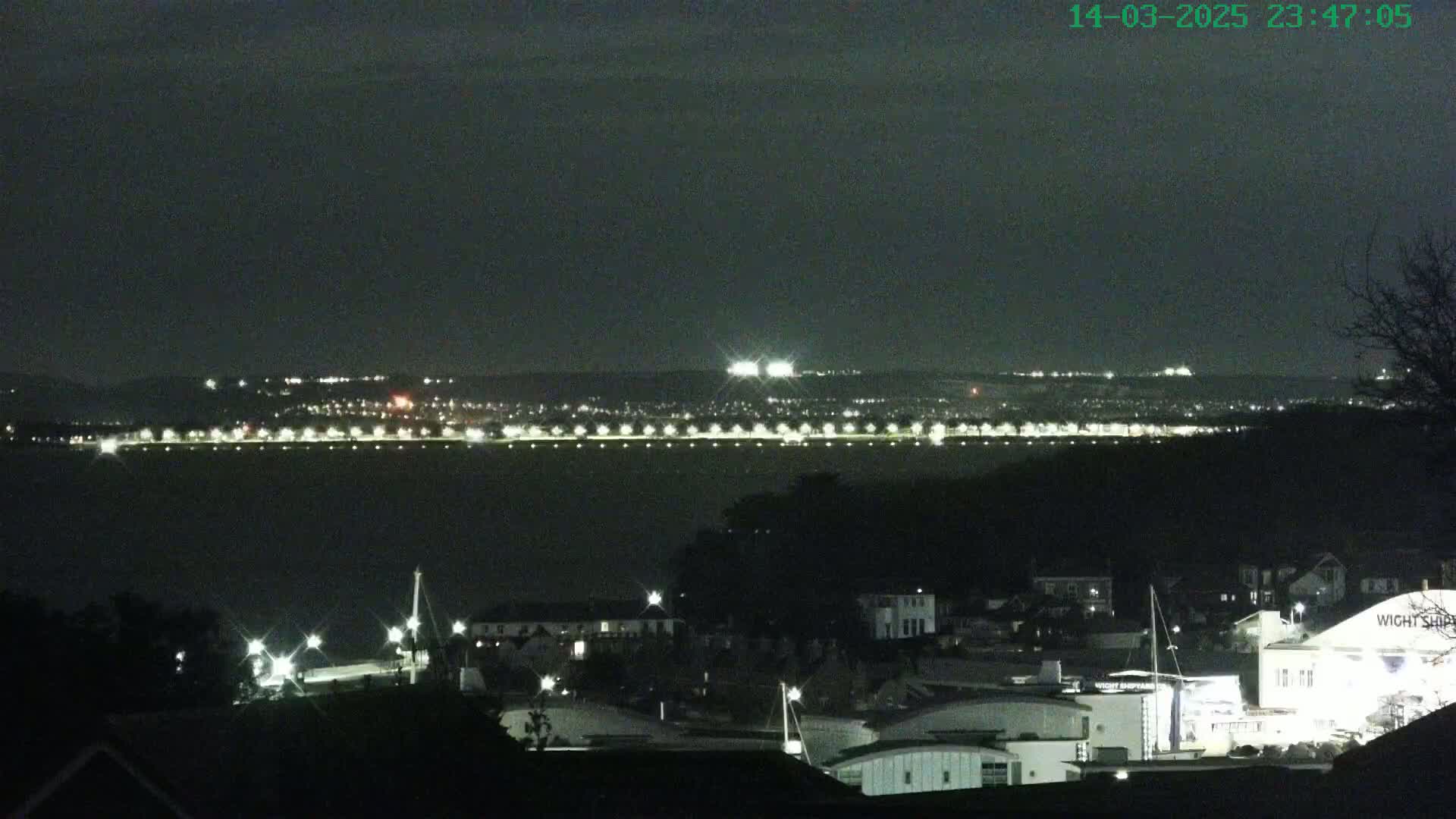 A nighttime view of a town's waterfront, illuminated by lights along a distant shoreline and buildings closer to the viewer, under a dark sky.