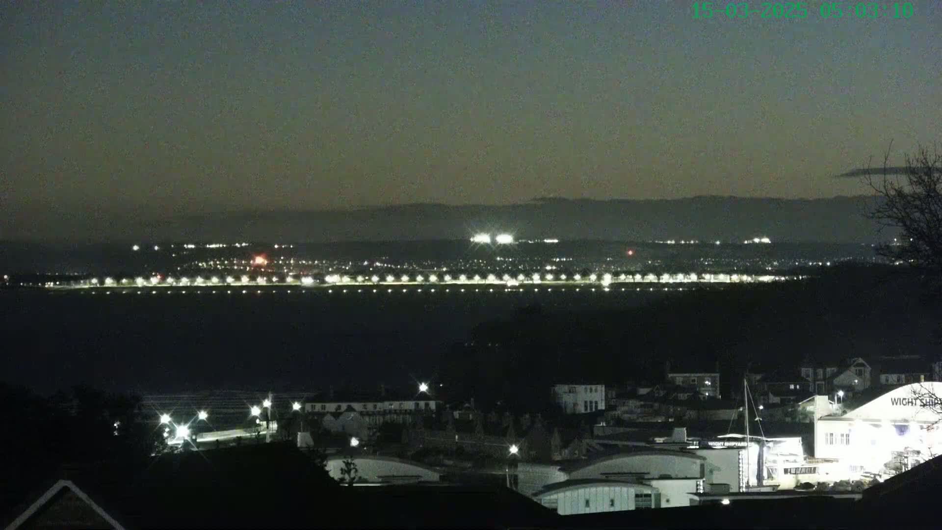 A nighttime, long-distance view of a city's illuminated waterfront,  backed by a dark, hilly landscape under a mostly clear sky.
