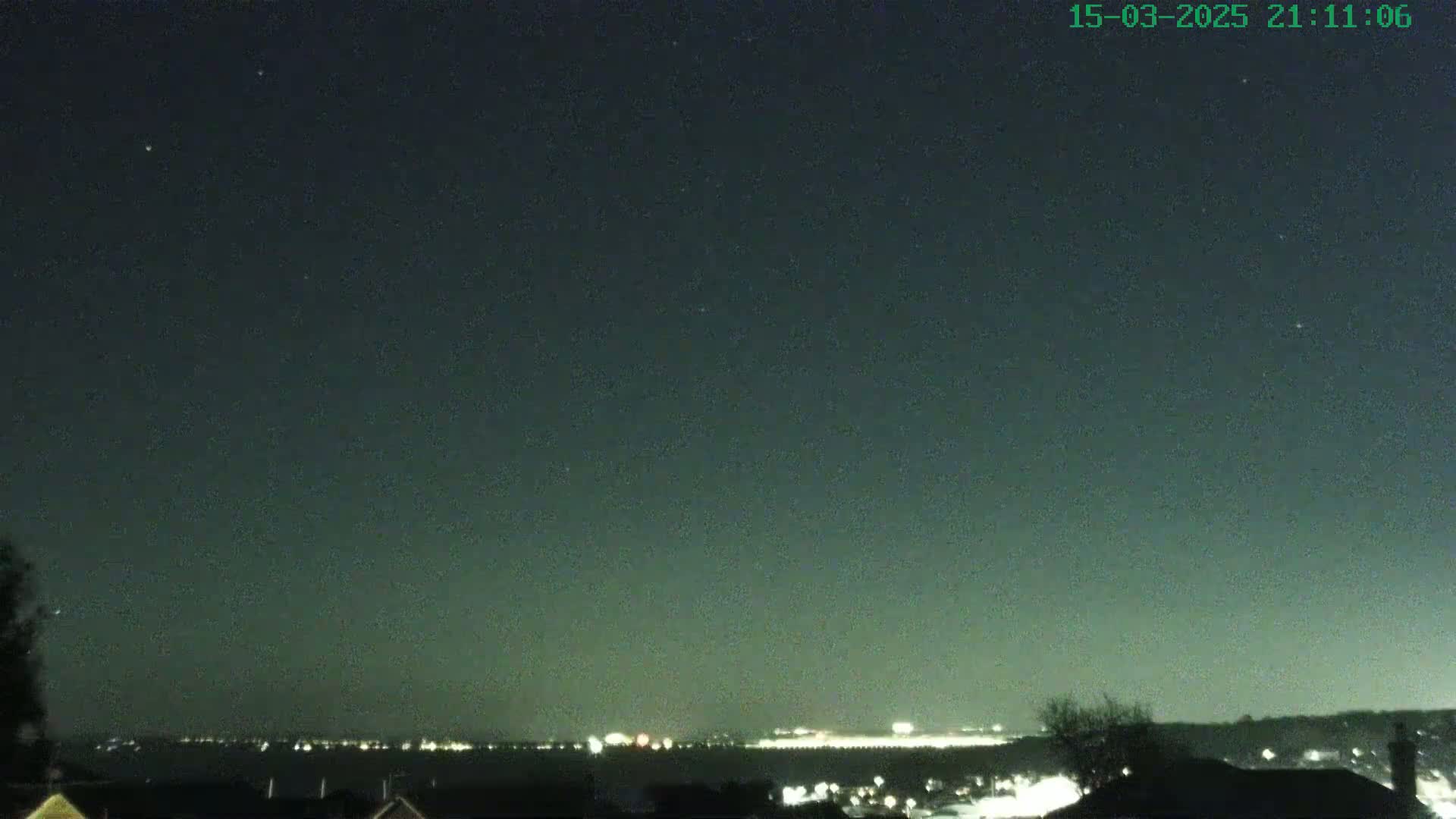 A nighttime view shows a dark sky with some stars above a distant, faintly lit cityscape across a body of water.