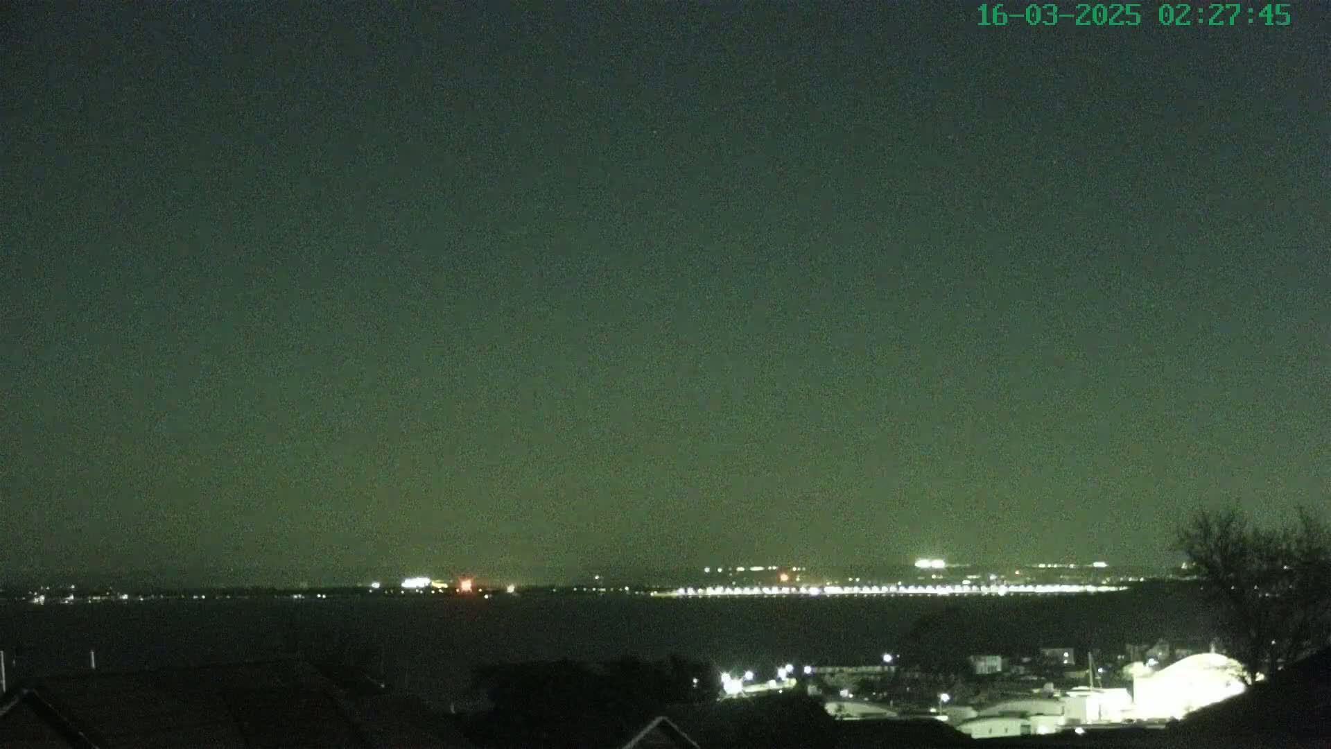 A nighttime view shows a dark body of water with a distant, faintly lit cityscape under a dark, clear sky.