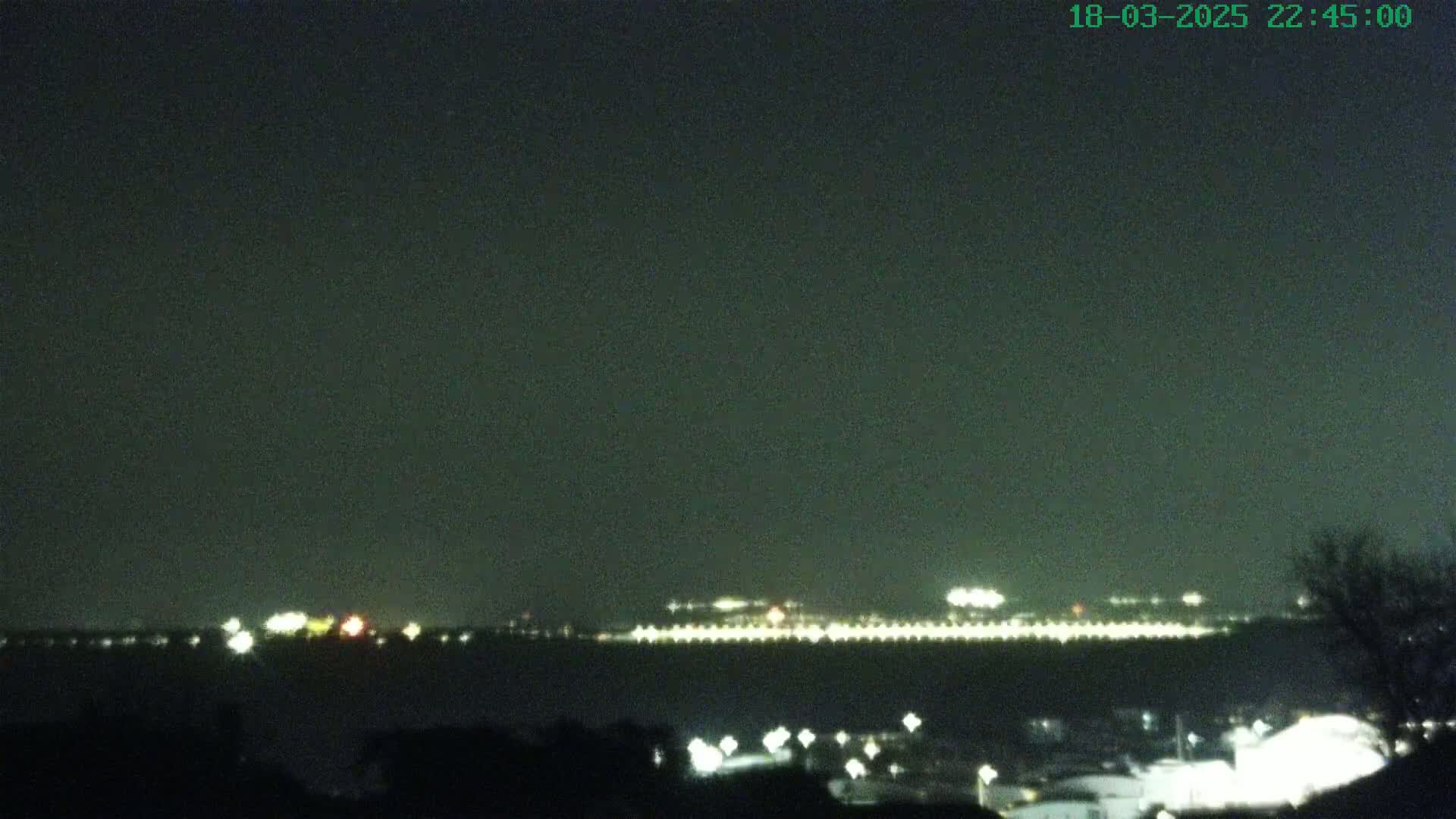 A nighttime long shot shows a distant city skyline illuminated with lights across a dark body of water under a clear, dark sky.