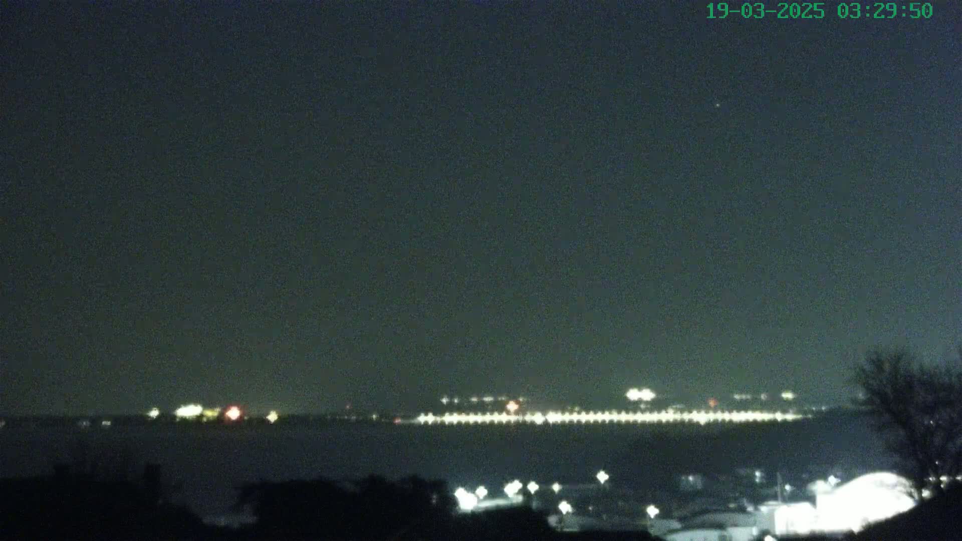 A nighttime long shot shows a line of distant lights across a body of water, with a few closer lights and dark silhouettes of trees and buildings in the foreground under a clear, dark sky.