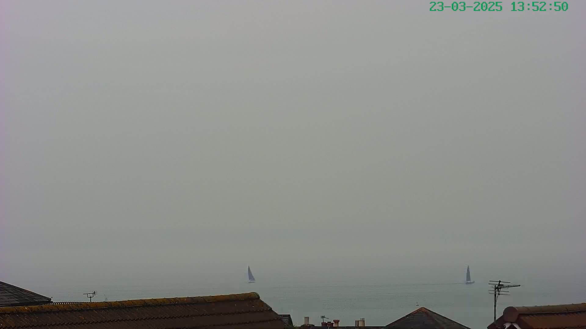 Two sailboats are visible on a hazy, gray sea, viewed from behind a row of house rooftops.