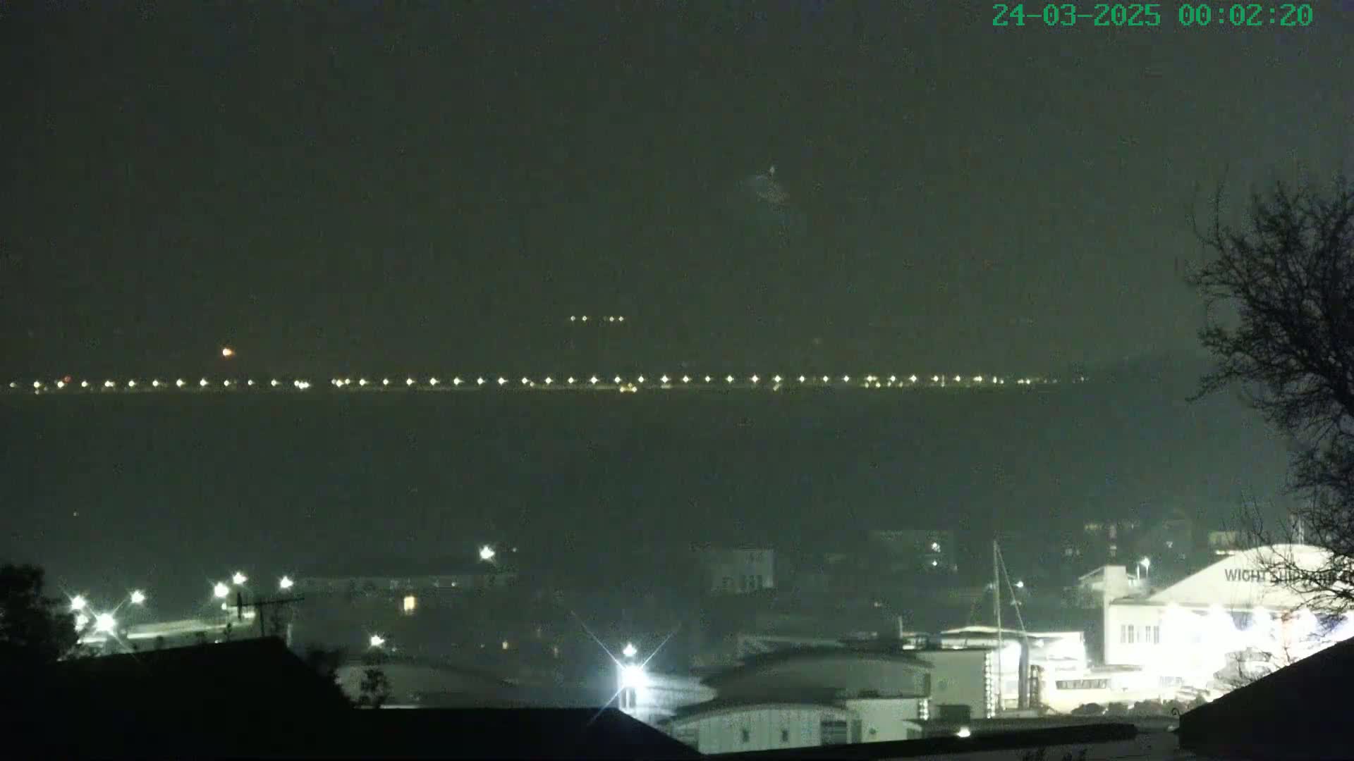 A nighttime view shows a line of lights across a body of water, with a hazy or foggy atmosphere, and a town or harbor with buildings and lights in the foreground.