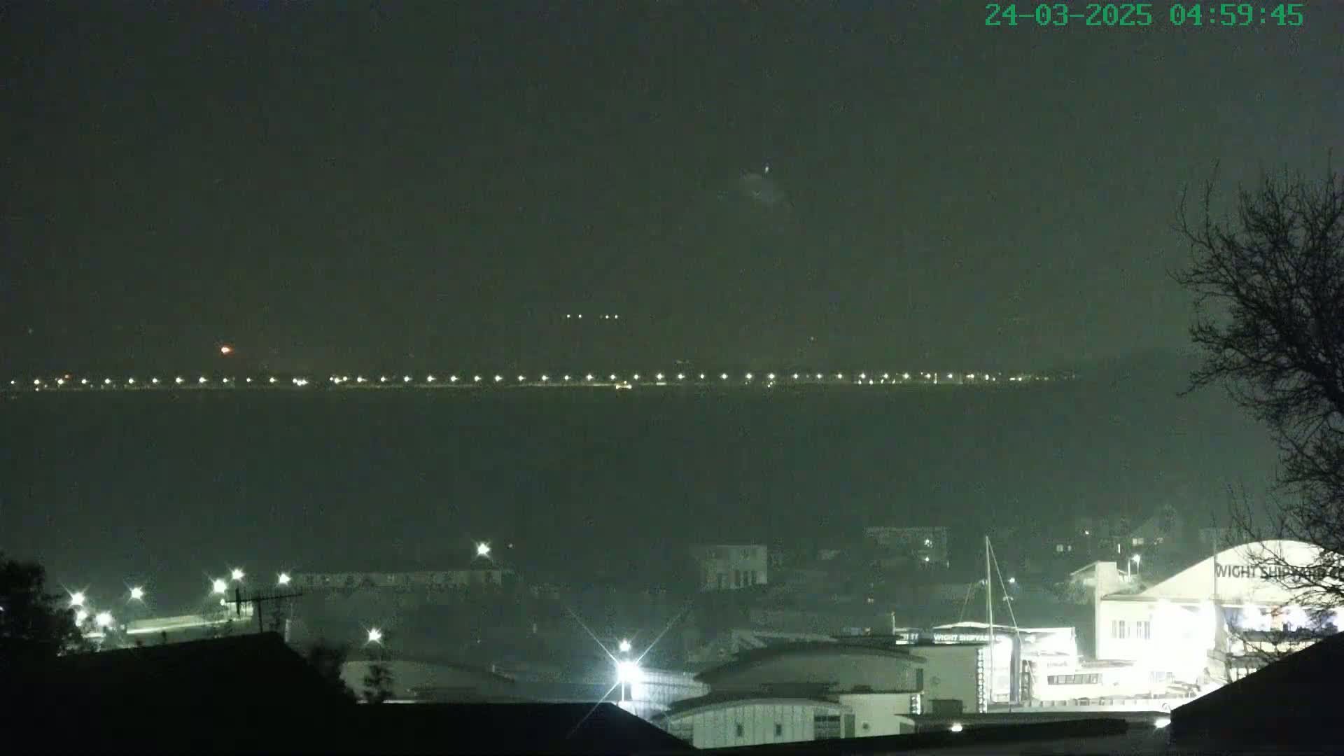 A hazy night scene shows a distant line of lights across a body of water, with a closer view of illuminated buildings and a marina.