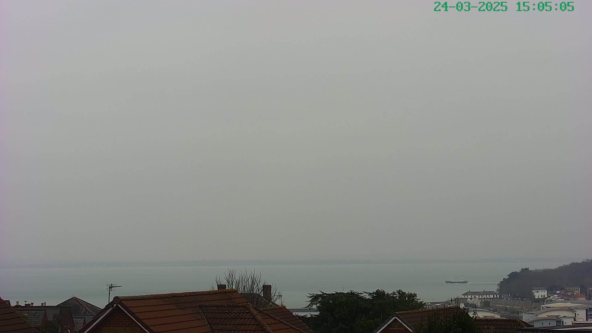 A hazy view of a calm body of water stretches to the horizon, beyond a row of brown-roofed houses and some trees.