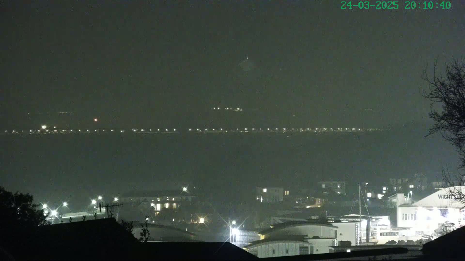 A hazy night scene shows a distant line of lights across a body of water, with a closer collection of illuminated buildings and structures below.