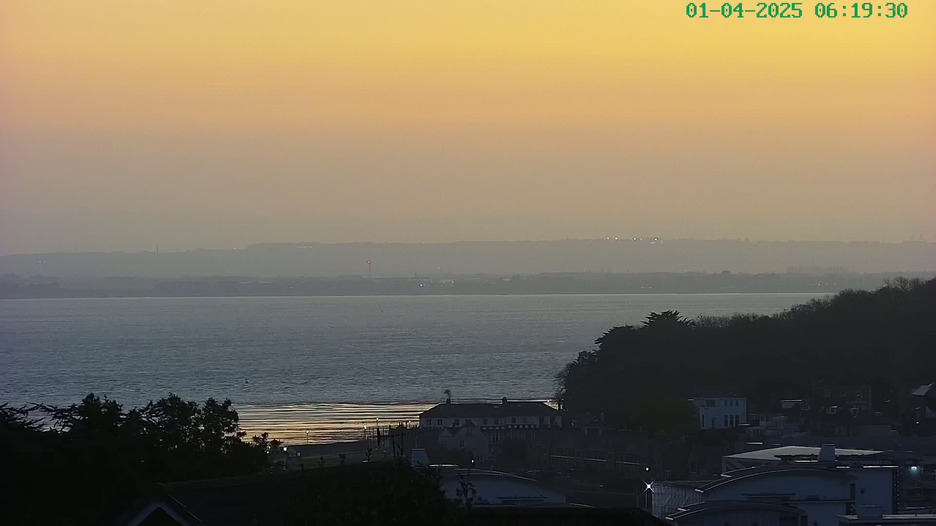 A hazy sunset over a calm body of water, with a beach and coastal buildings visible in the foreground.
