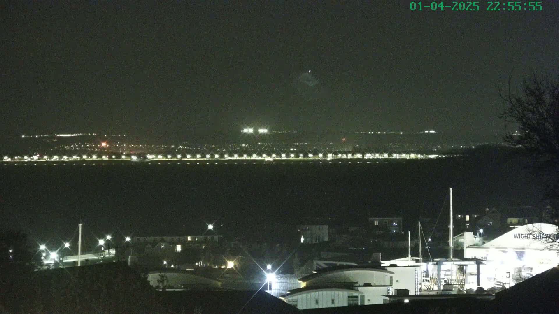 A nighttime long shot shows a dark body of water with a distant illuminated shoreline and a closer view of buildings and lights at a marina under a clear, dark sky.