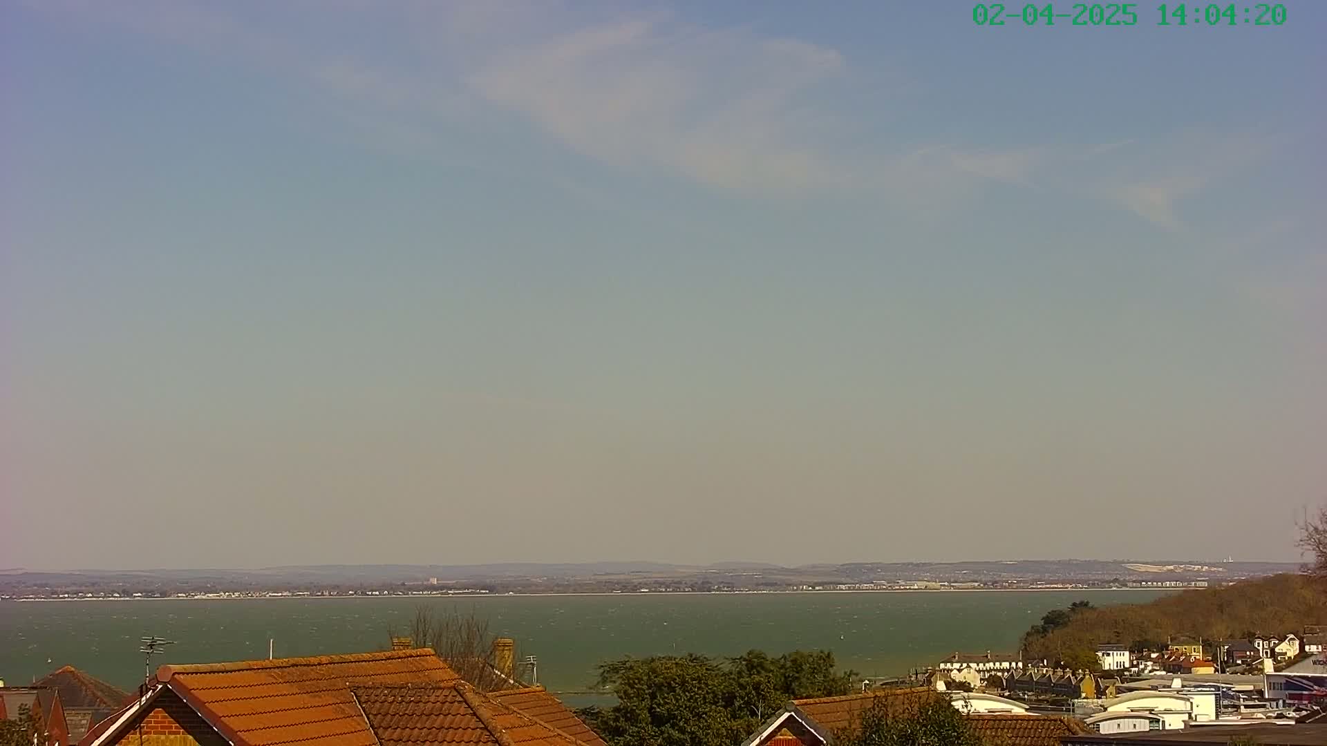 A slightly hazy, sunny day overlooks a calm body of water with a distant shoreline and rooftops of houses in the foreground.