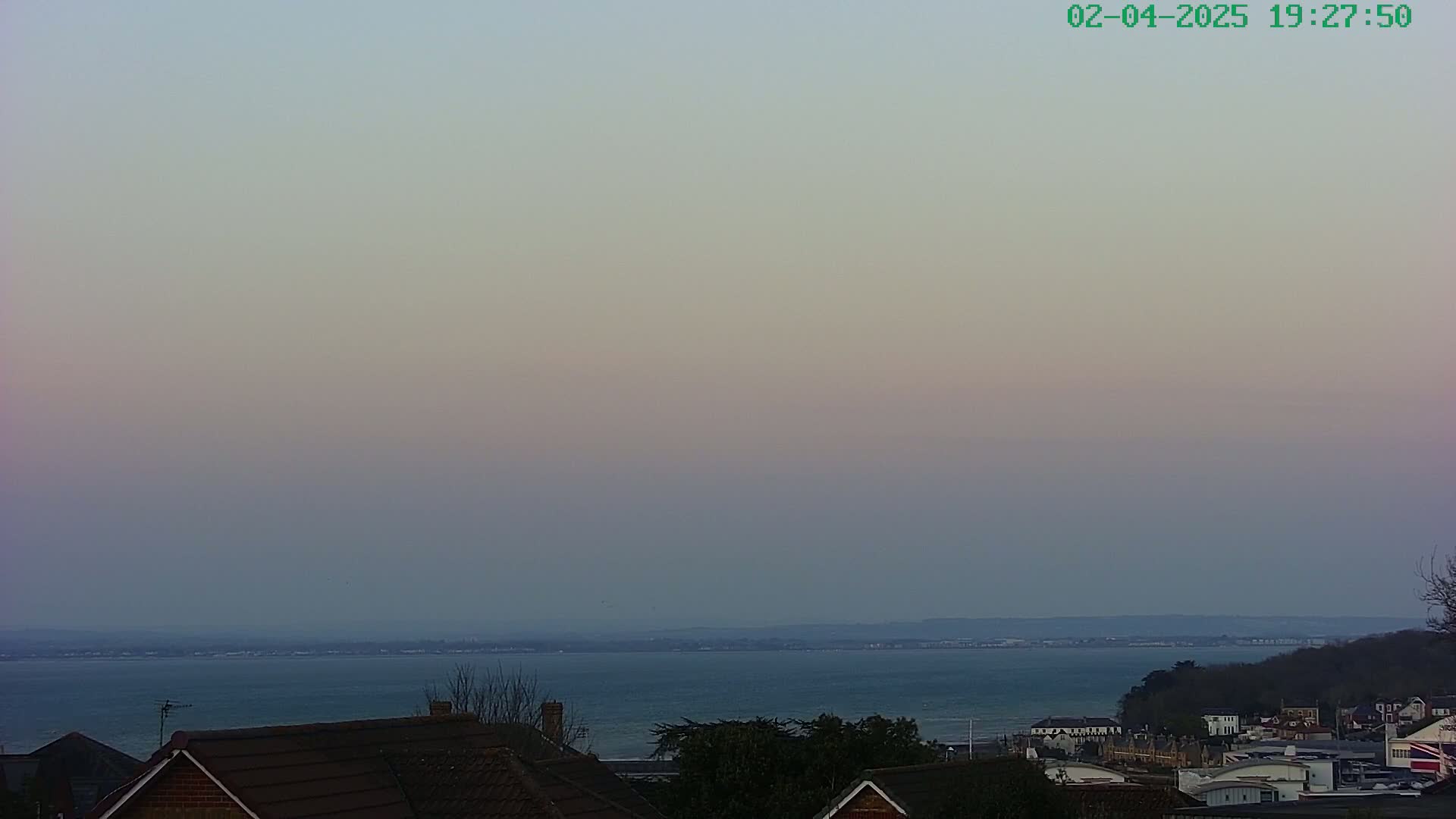 A calm body of water stretches to a distant shoreline under a pale, twilight sky, visible from an elevated viewpoint overlooking residential rooftops.