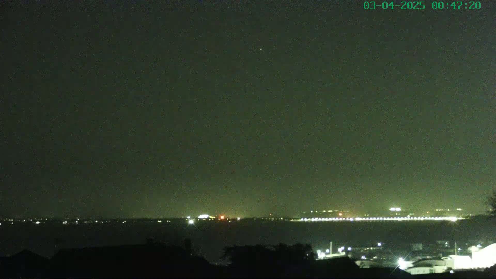 A nighttime long shot shows a body of water with a distant city skyline faintly illuminated, under a dark, clear sky.