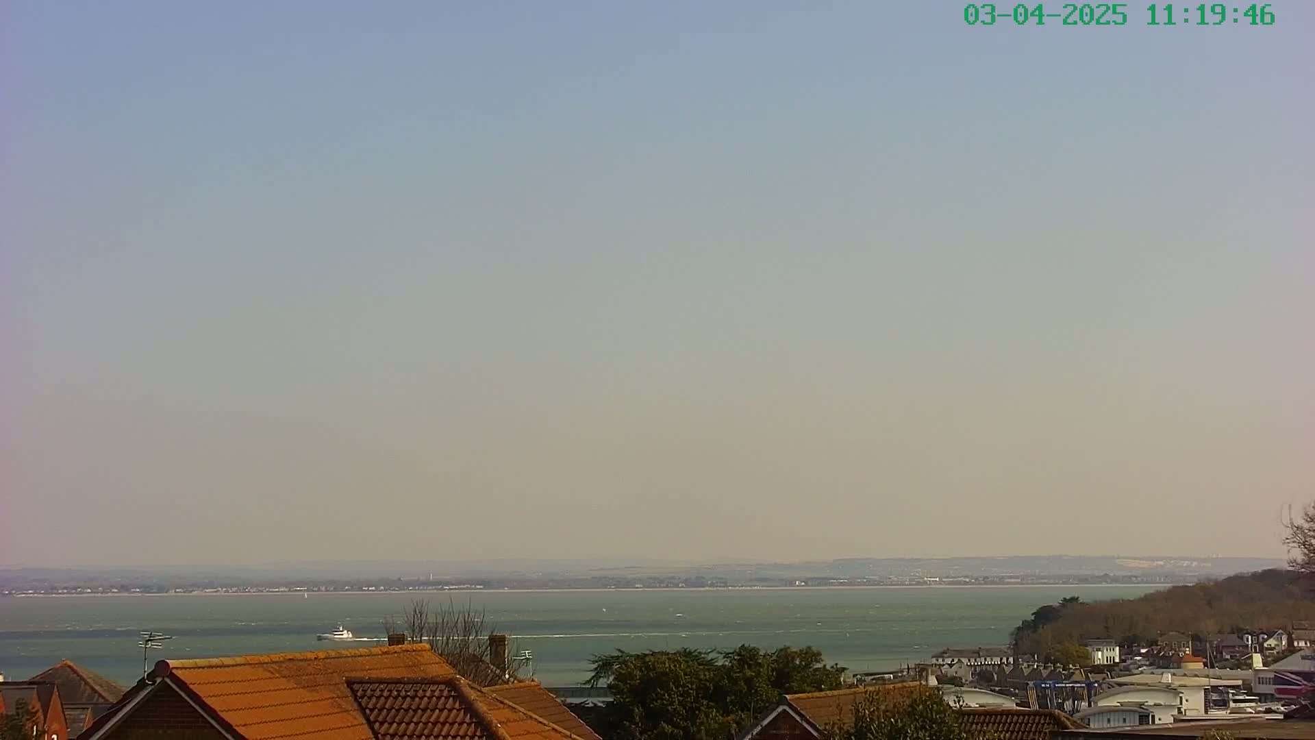 A hazy, sunny day overlooks a calm body of water with a distant shoreline, seen from atop a collection of houses with orange roofs.