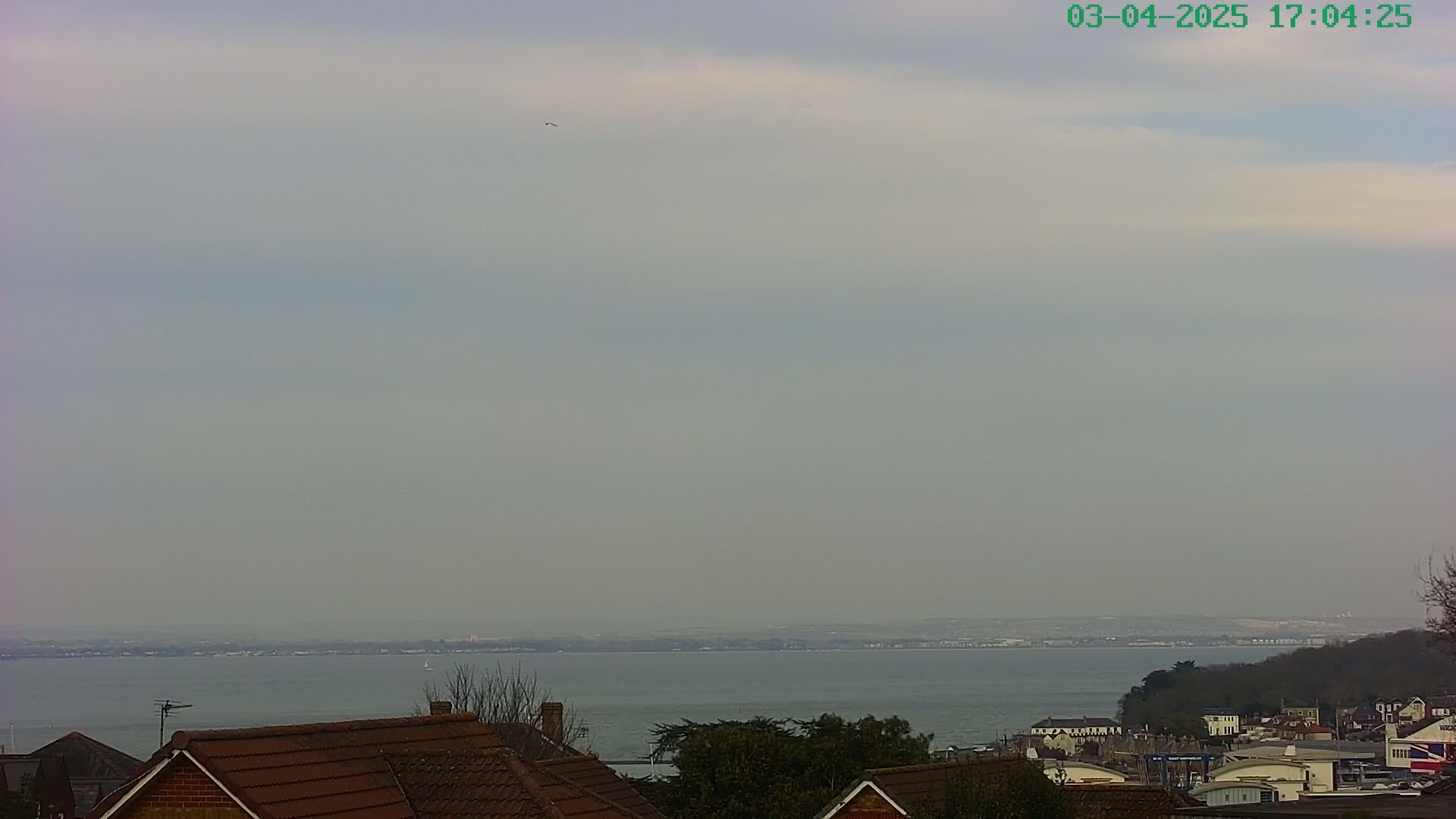 A hazy view shows a calm body of water extending to a distant shoreline under a mostly cloudy sky, with rooftops of houses and trees in the foreground.