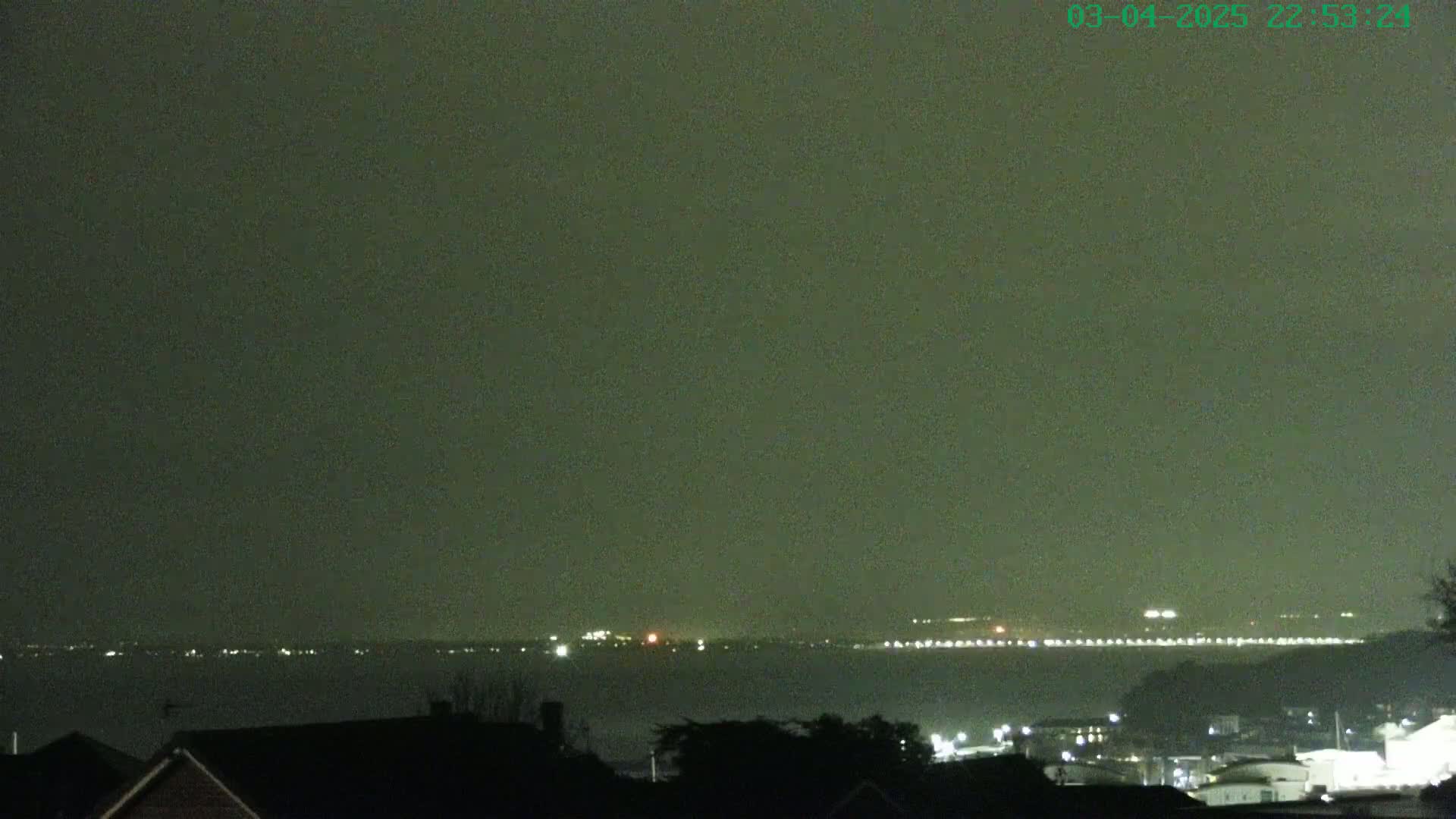 A nighttime view shows a body of water with distant lights across the horizon under a dark, overcast sky.
