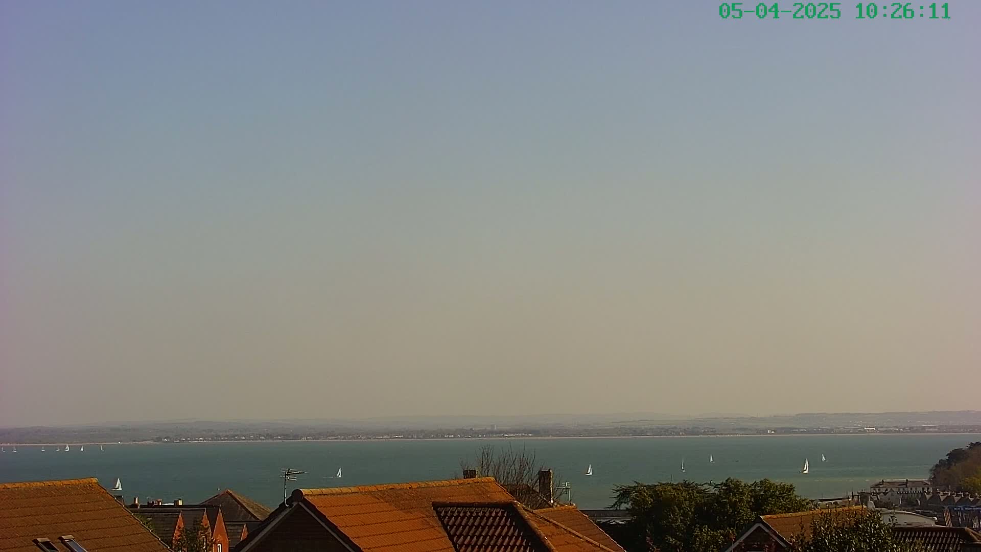 A hazy, sunny day reveals a calm body of water dotted with sailboats, viewed from above a line of brown-roofed houses.
